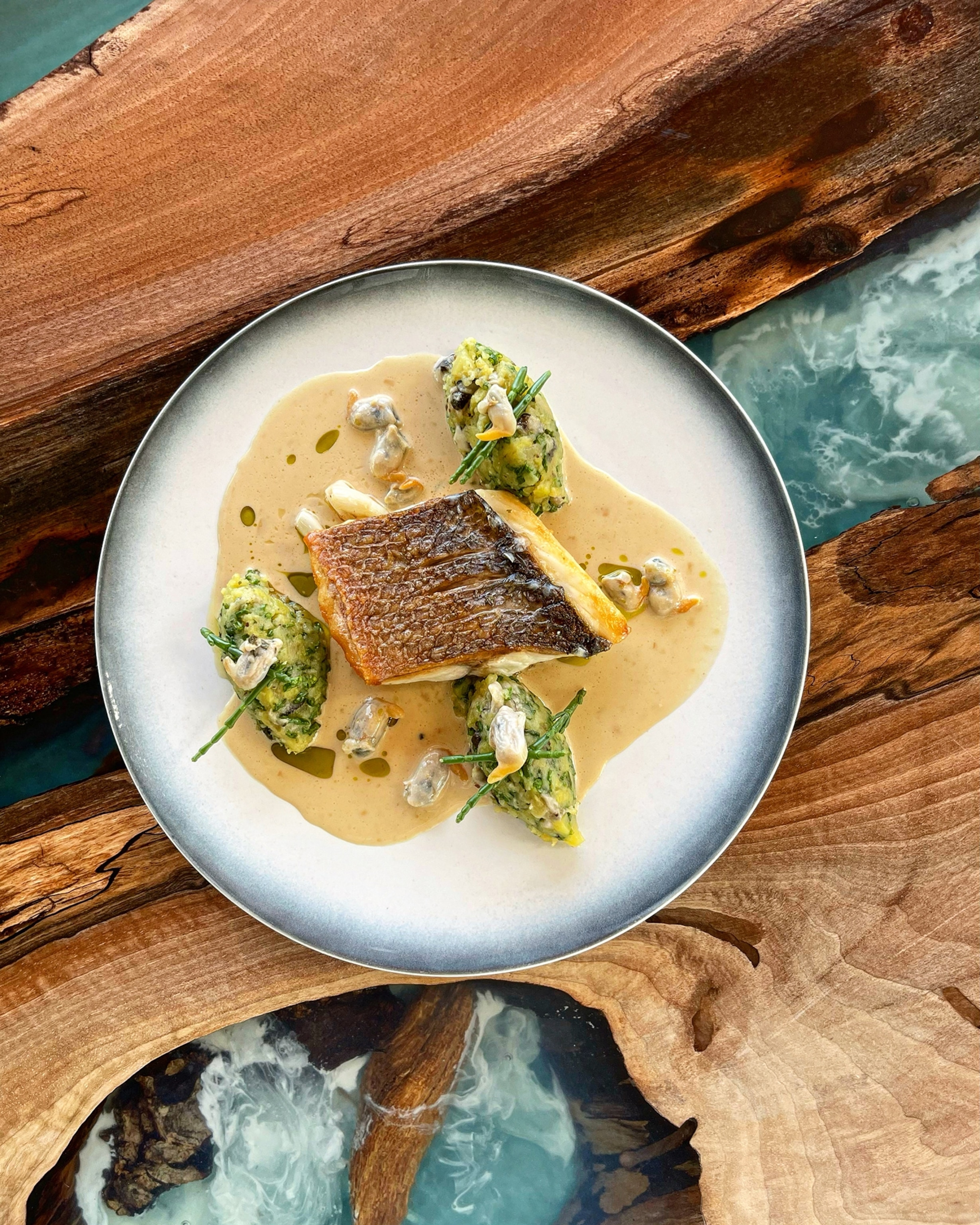 A plate of Pan seared Line caught Sea Bass, comfit cabbage, baby clams citronelle emulsion against a blue marbled and wood table.