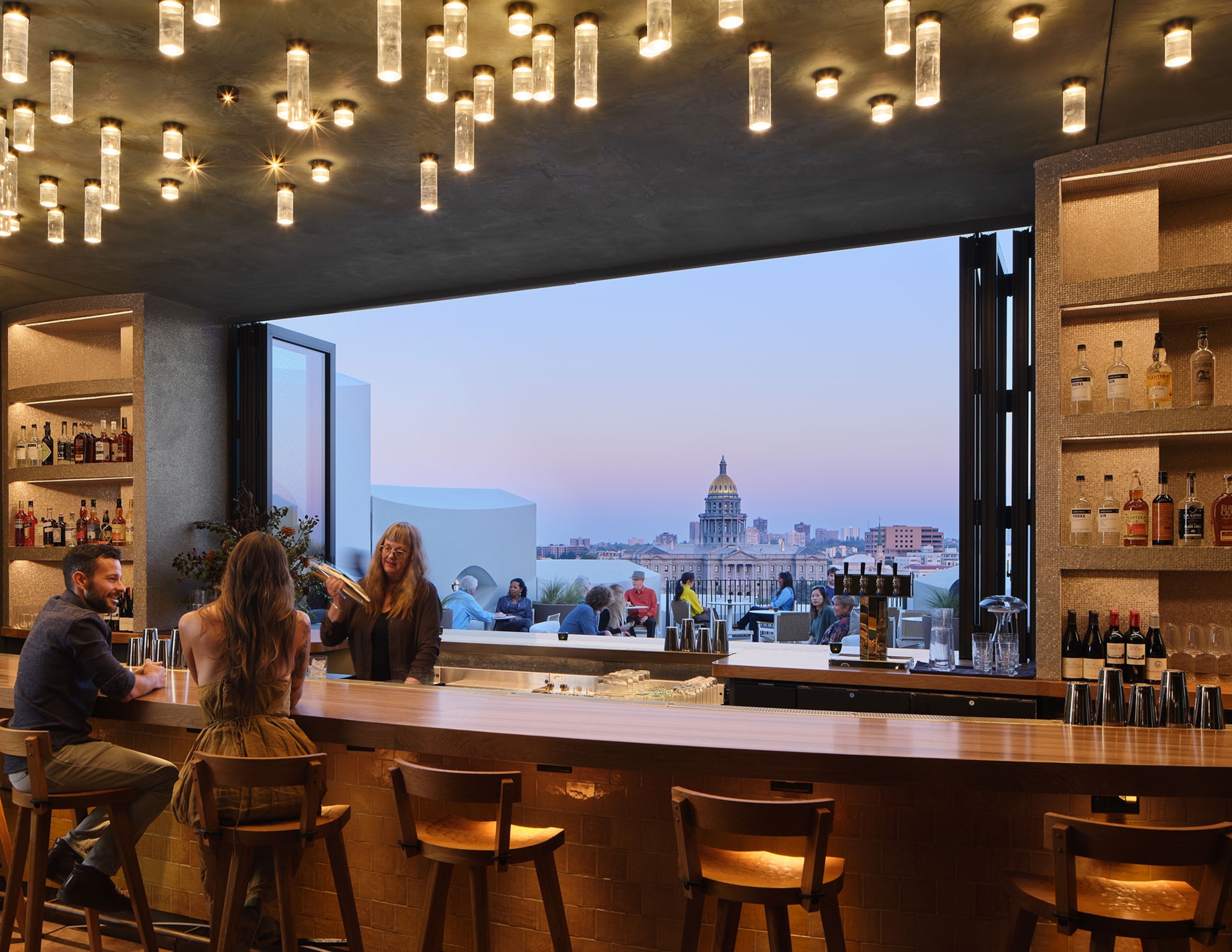 People sitting at a bar with a big open window looking out onto a city with the sun setting in the background.