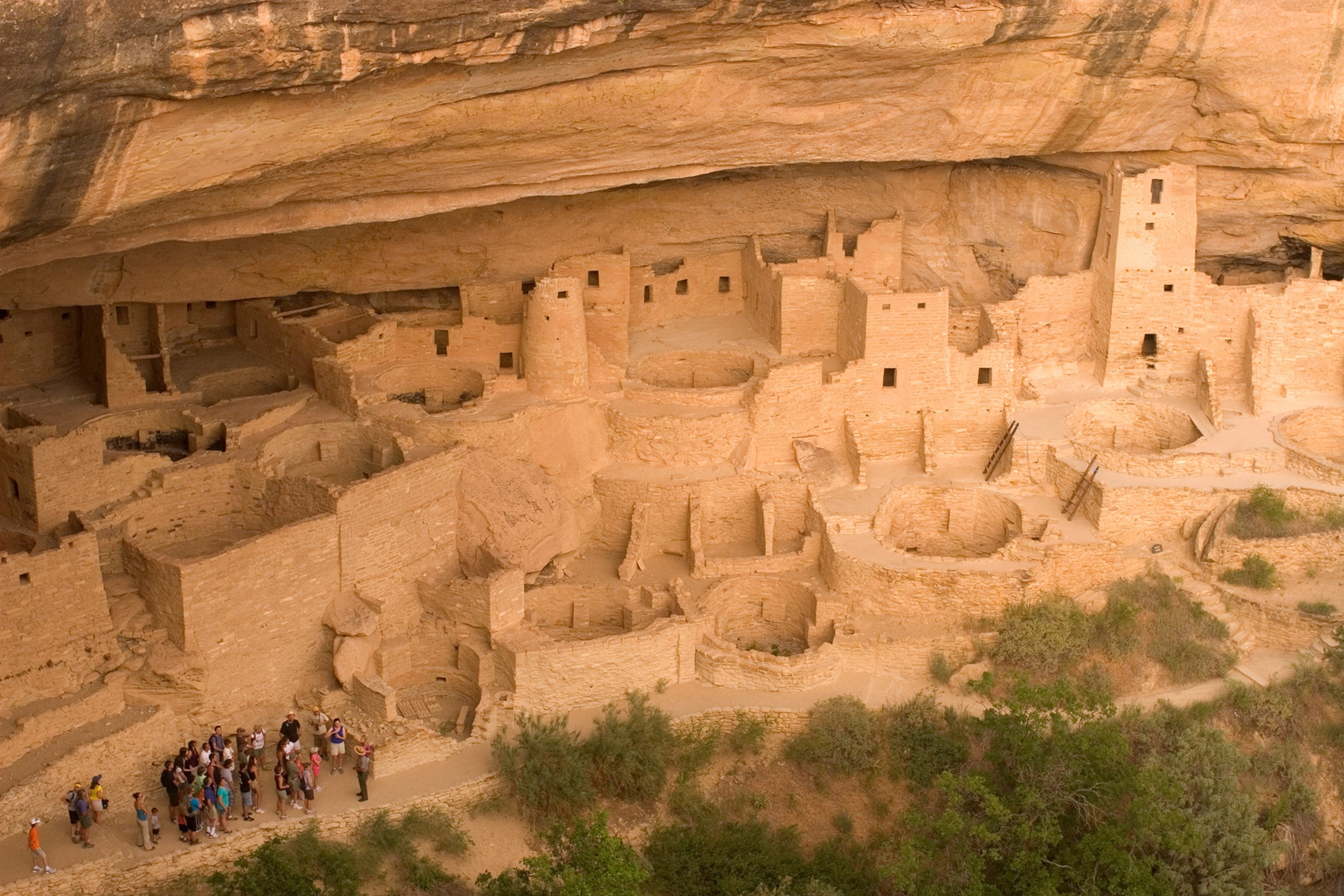 A group of people, tour old structures built into cliff.