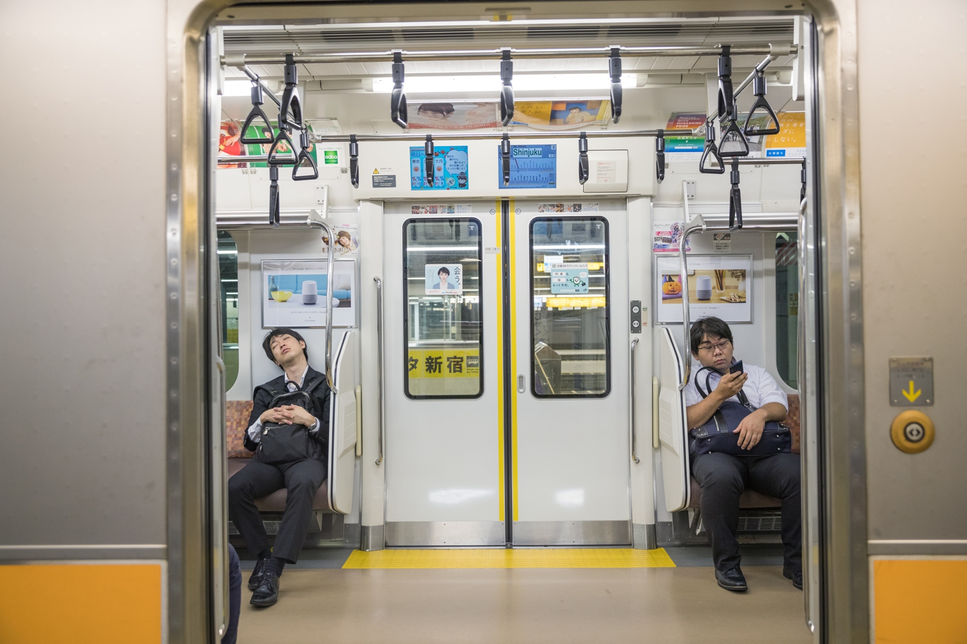 one man napping and one man on his phone on a subway train with the doors open