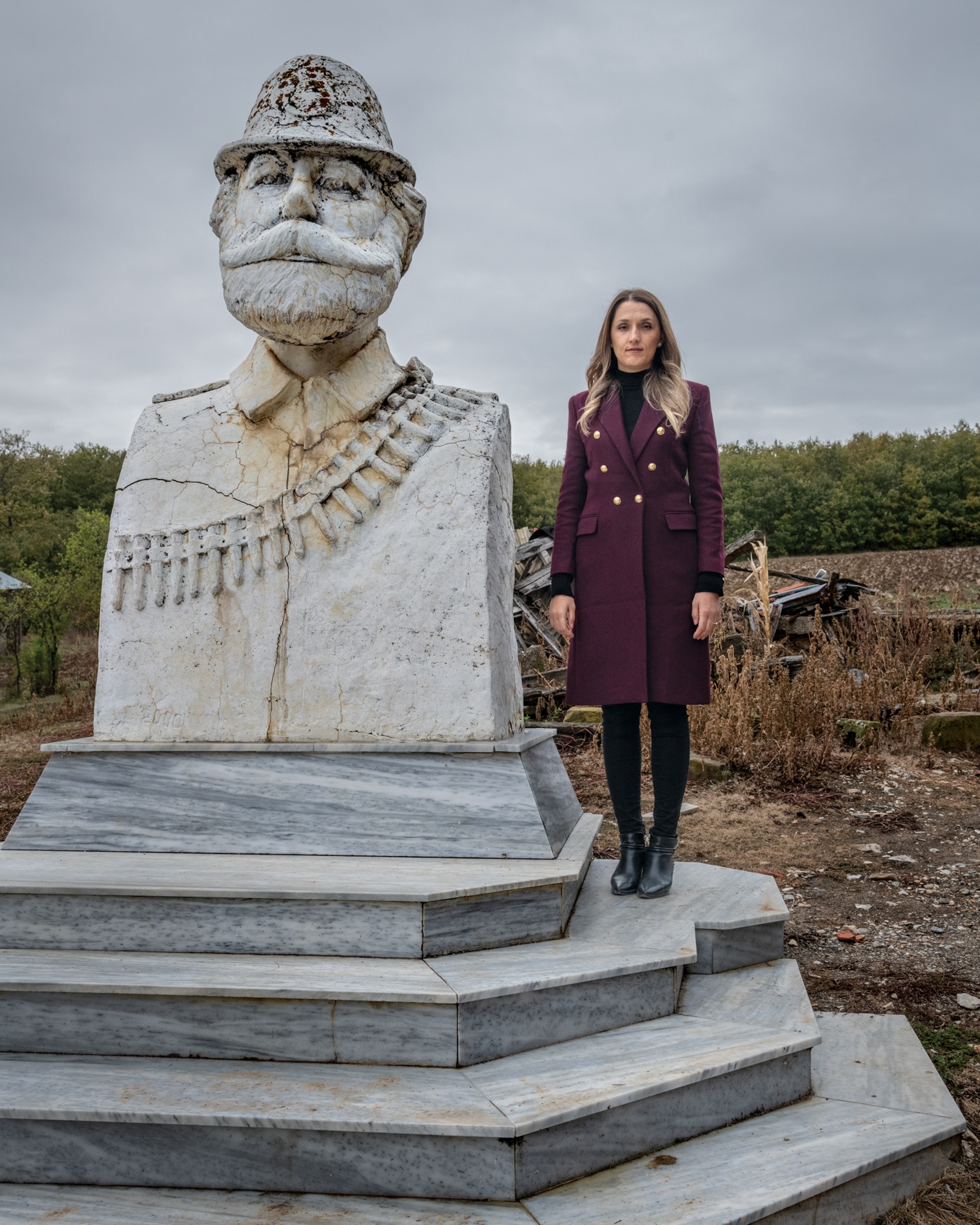 Picture of a woman standing next to a very large statue of a mans torso made of stone wearing a had, ammunition over his shoulder, and has a mustache.
