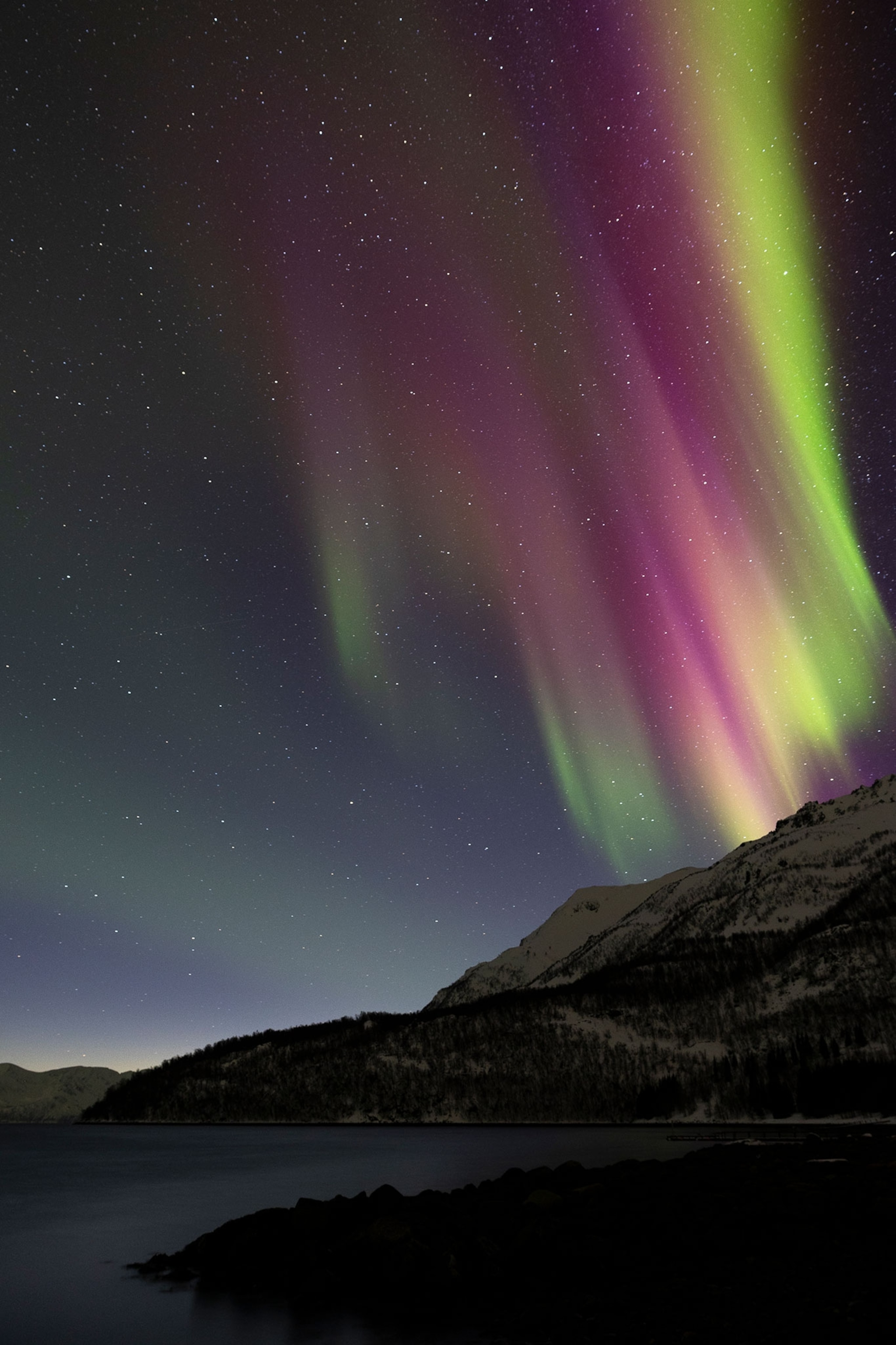 The Northern Lights fill a night sky above a mountain range and body of water below.