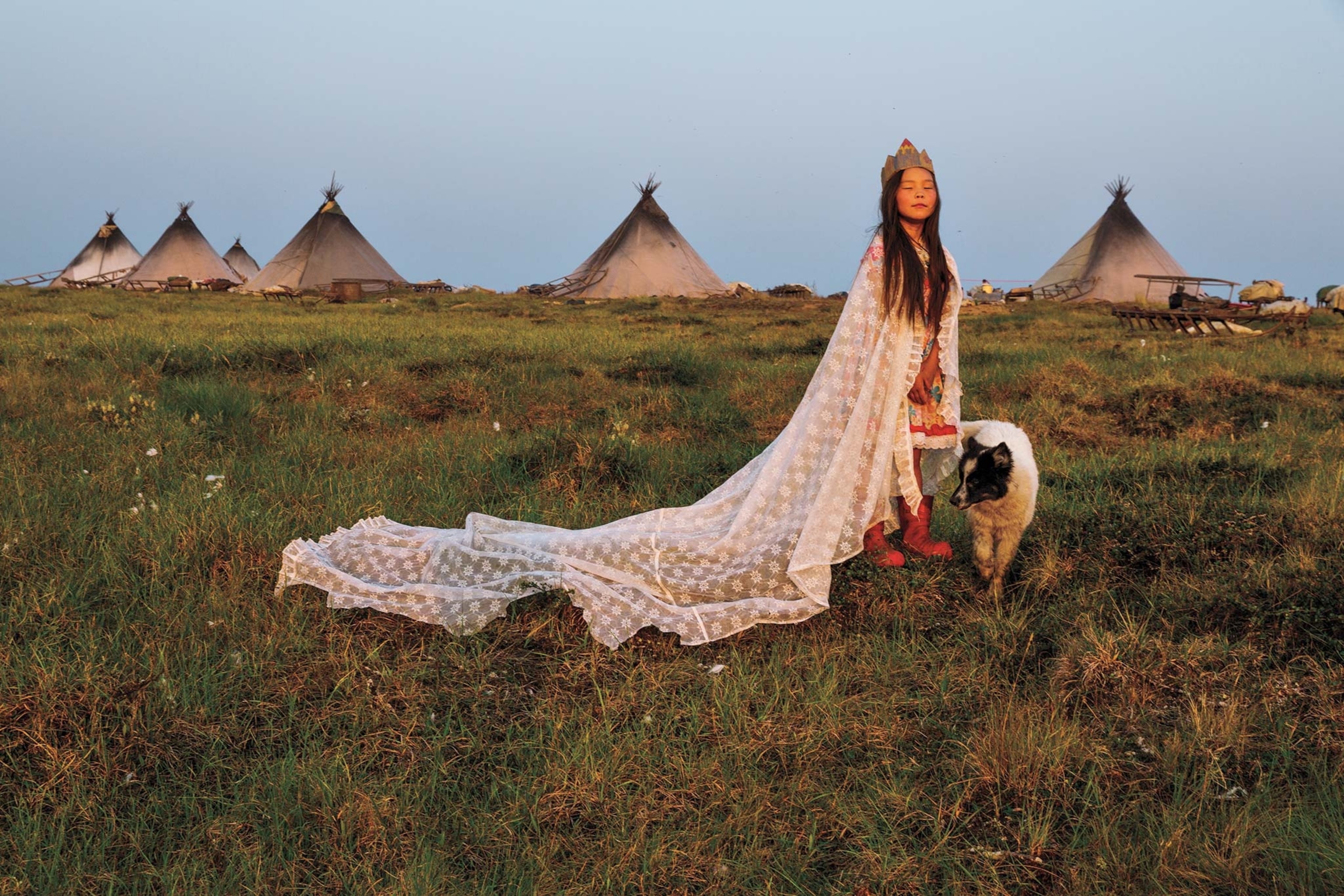 A woman wears a lace curtain as a robe and a cardboard crown in front of an Indigenous camp