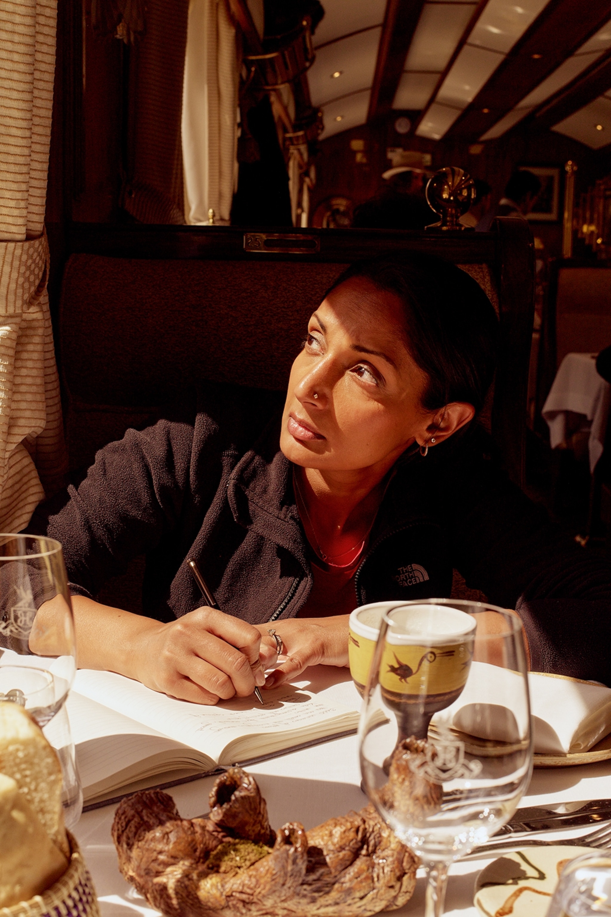 A female writer sitting at a set table with a notebook and pen, looking out the moving train window.