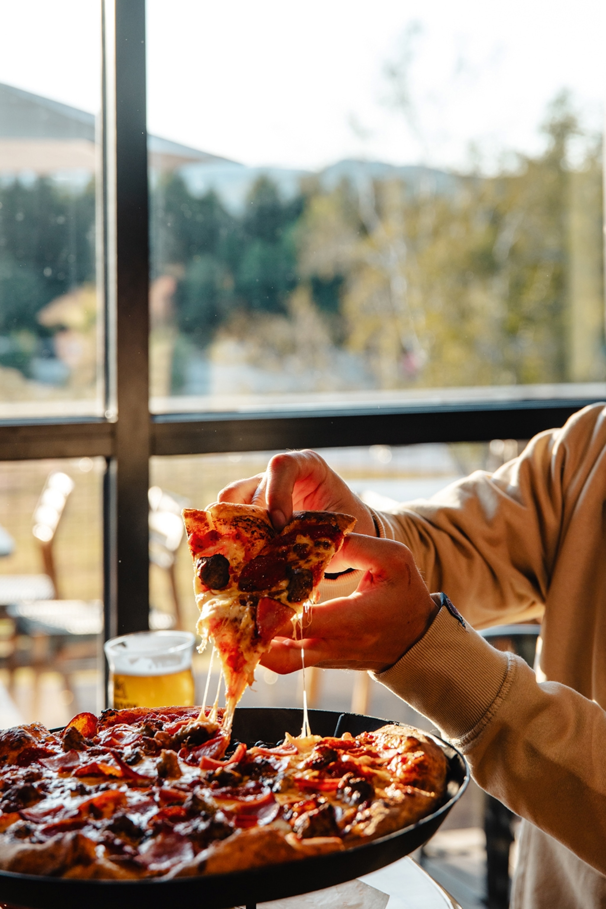A person picking up a slice of cheesy pizza off the plate by a floor-length window.