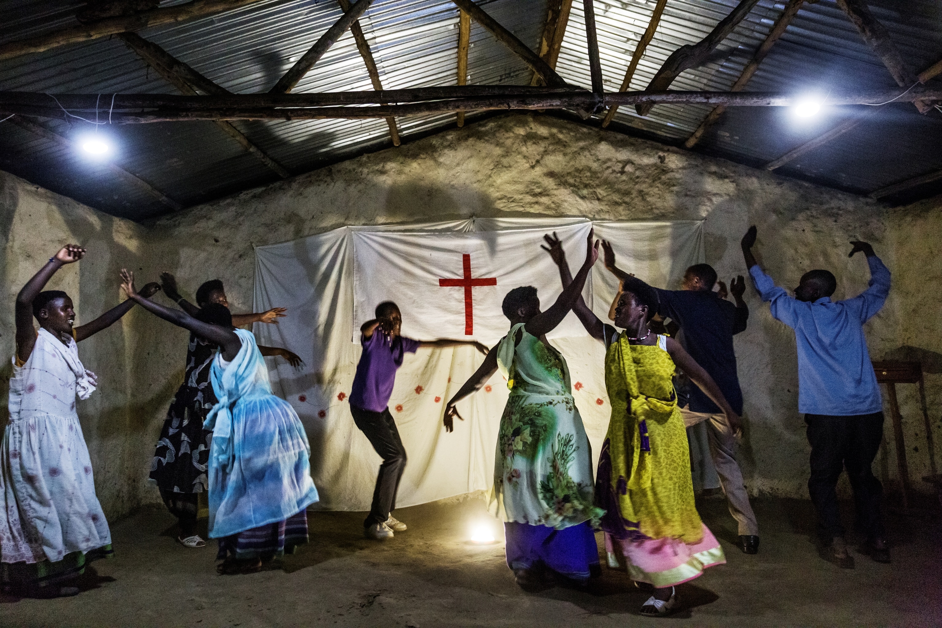 Ten Black women and men in colorful garb dance inside a small, rustic church with a corrugated tin ceiling. On the stone wall behind them hangs a large white textile featuring a red cross appliqué.