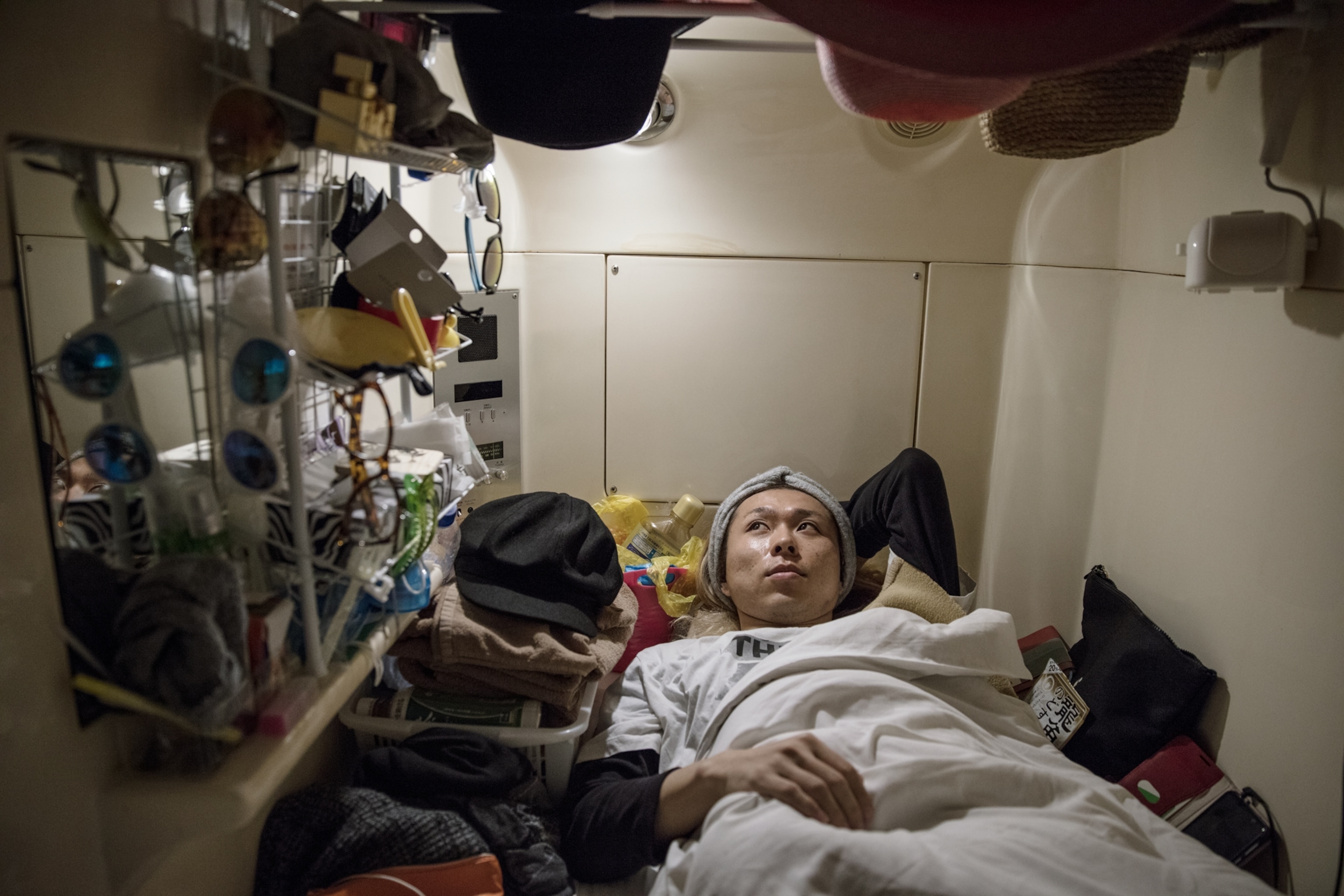 a man resting in his capsule bedroom with many personal items within his small bedroom