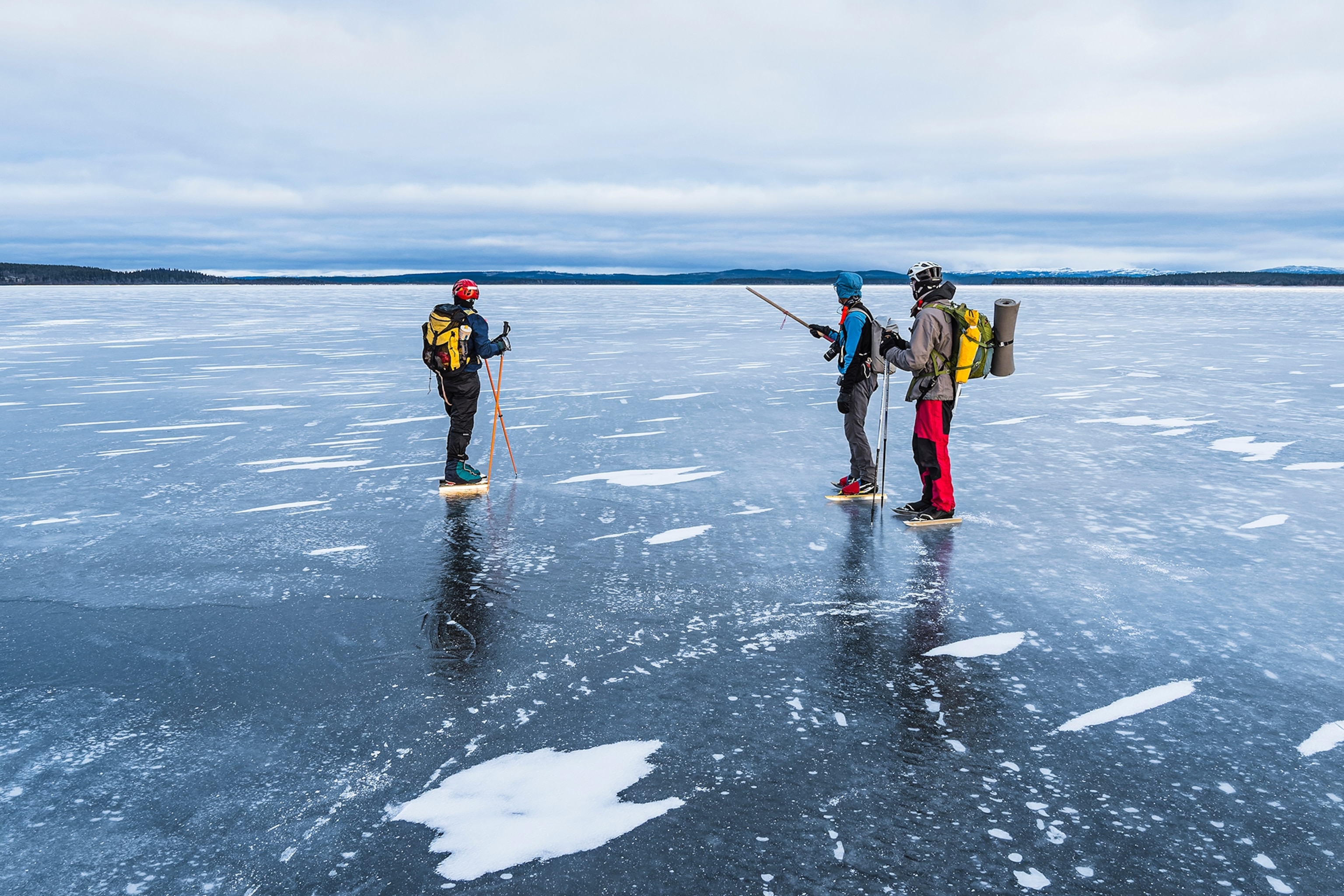 Three skaters in thermal hiking gear on a wide frozen lake.