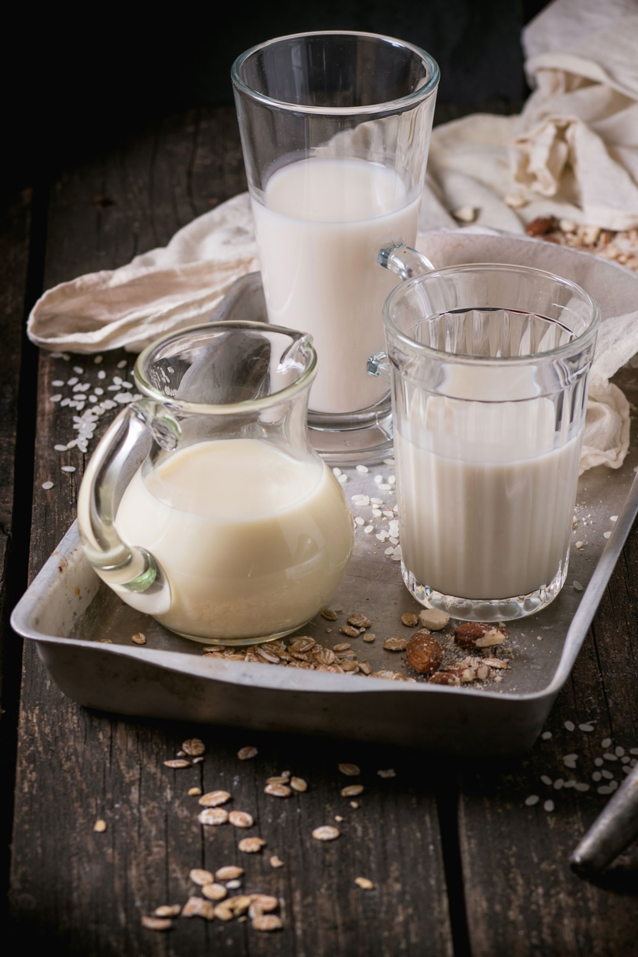 Set of non-dairy milk in glass cups and jug on old aluminum tray with rice grains, oat flakes and almond over old wooden table.