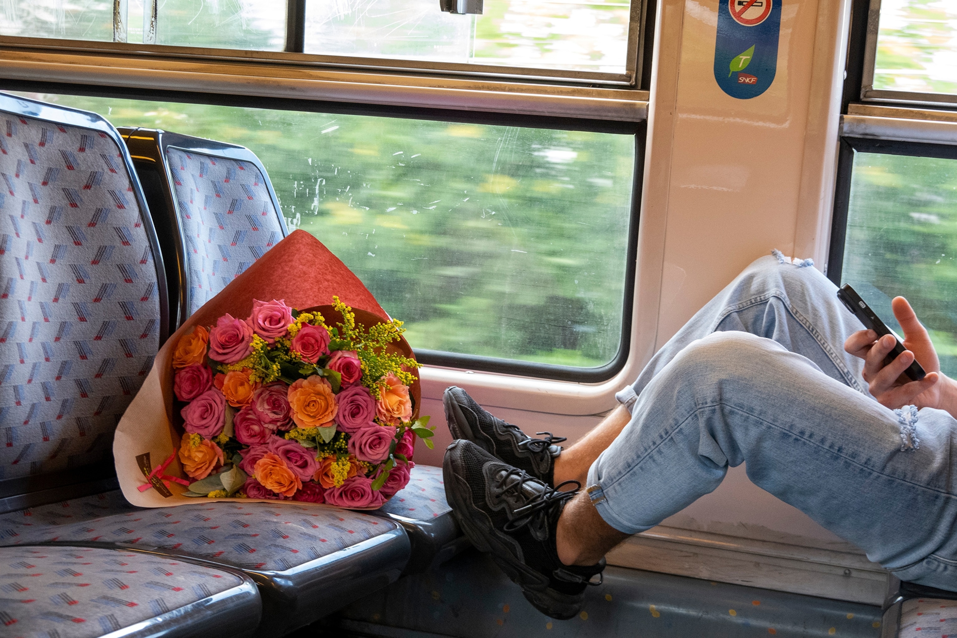 A relaxed young passenger checks his cell phone on the train. A large bouquet of roses rests on the seat in front of him.