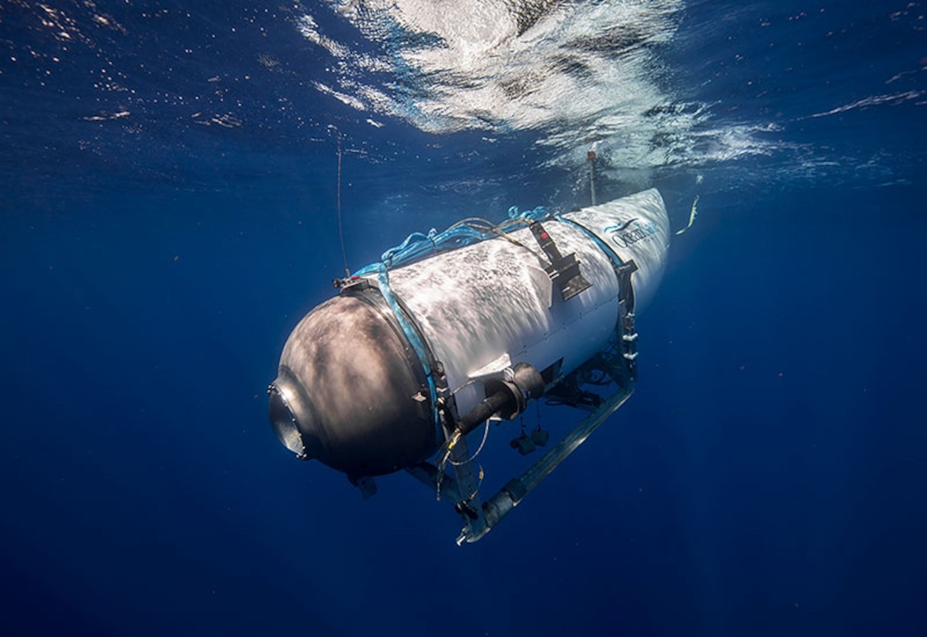 A white and grey submarine decends in deep blue water with lots of light coming through near the surface.
