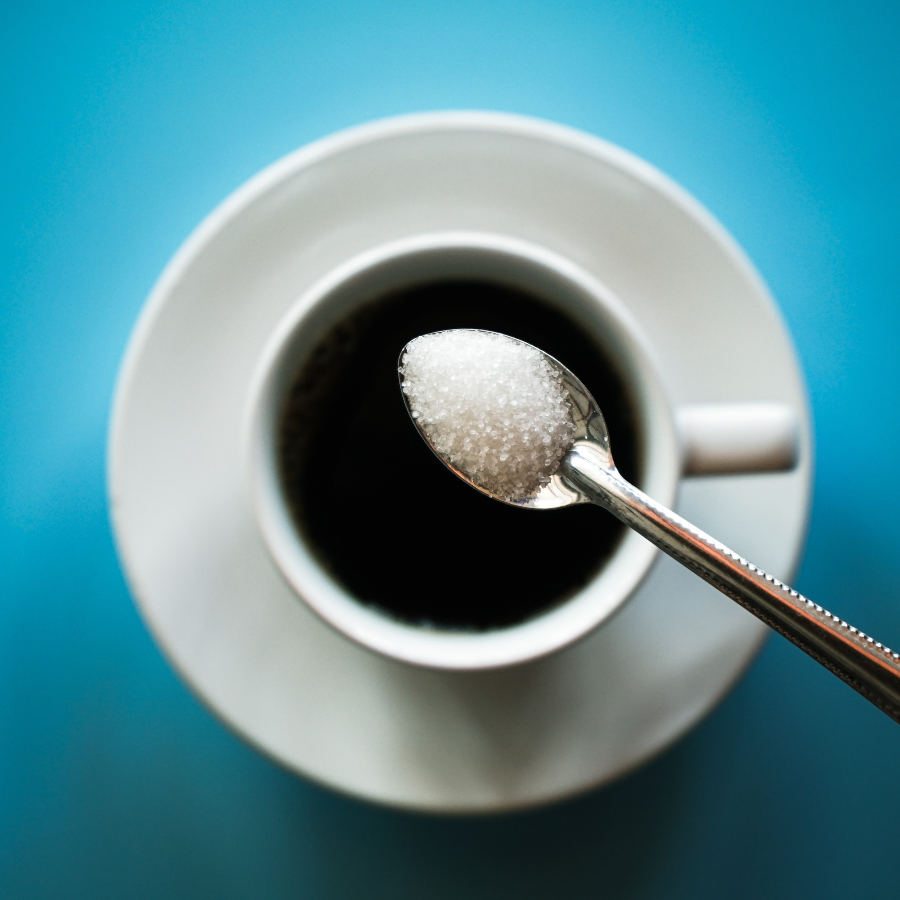 Top view of a spoonful of white sugar over a white cup of black coffee on a white saucer, against a blue background.