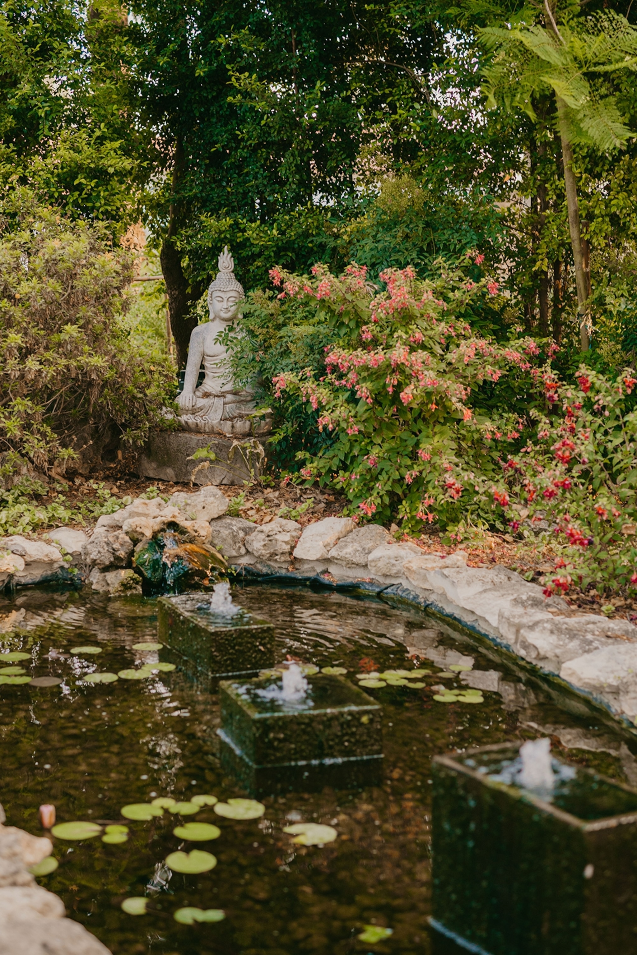 A serene pond surrounded by blooming bushed and vegetation, a buddha statue in the back.