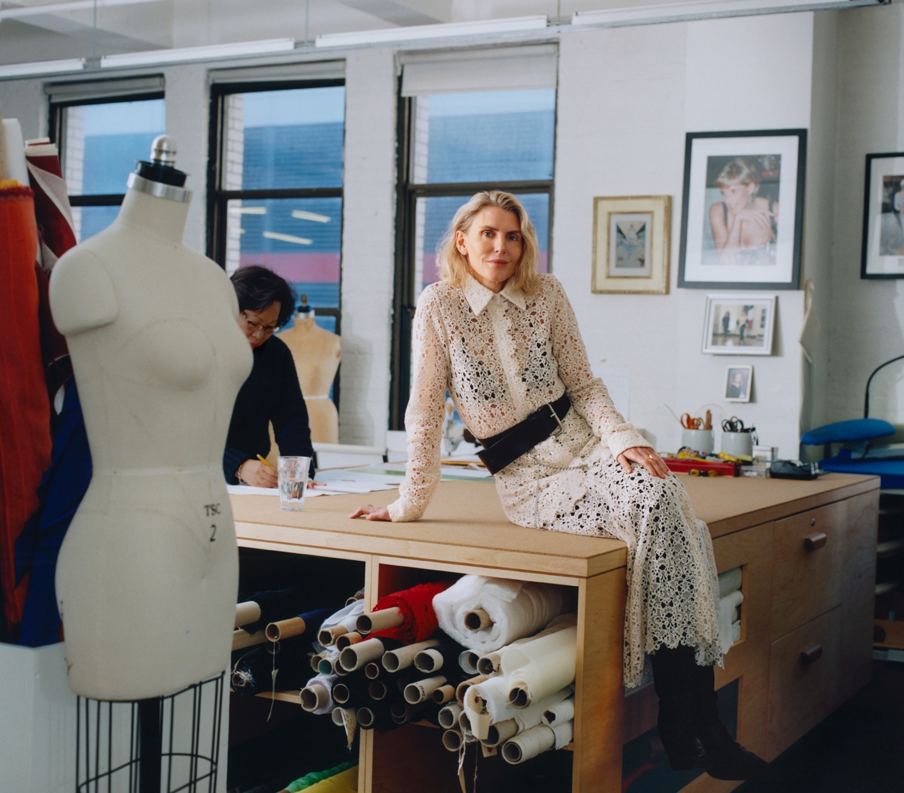 A woman in a white dress sits on top of a counter that has fabric stored underneath. There is a mannequin seen on the side.