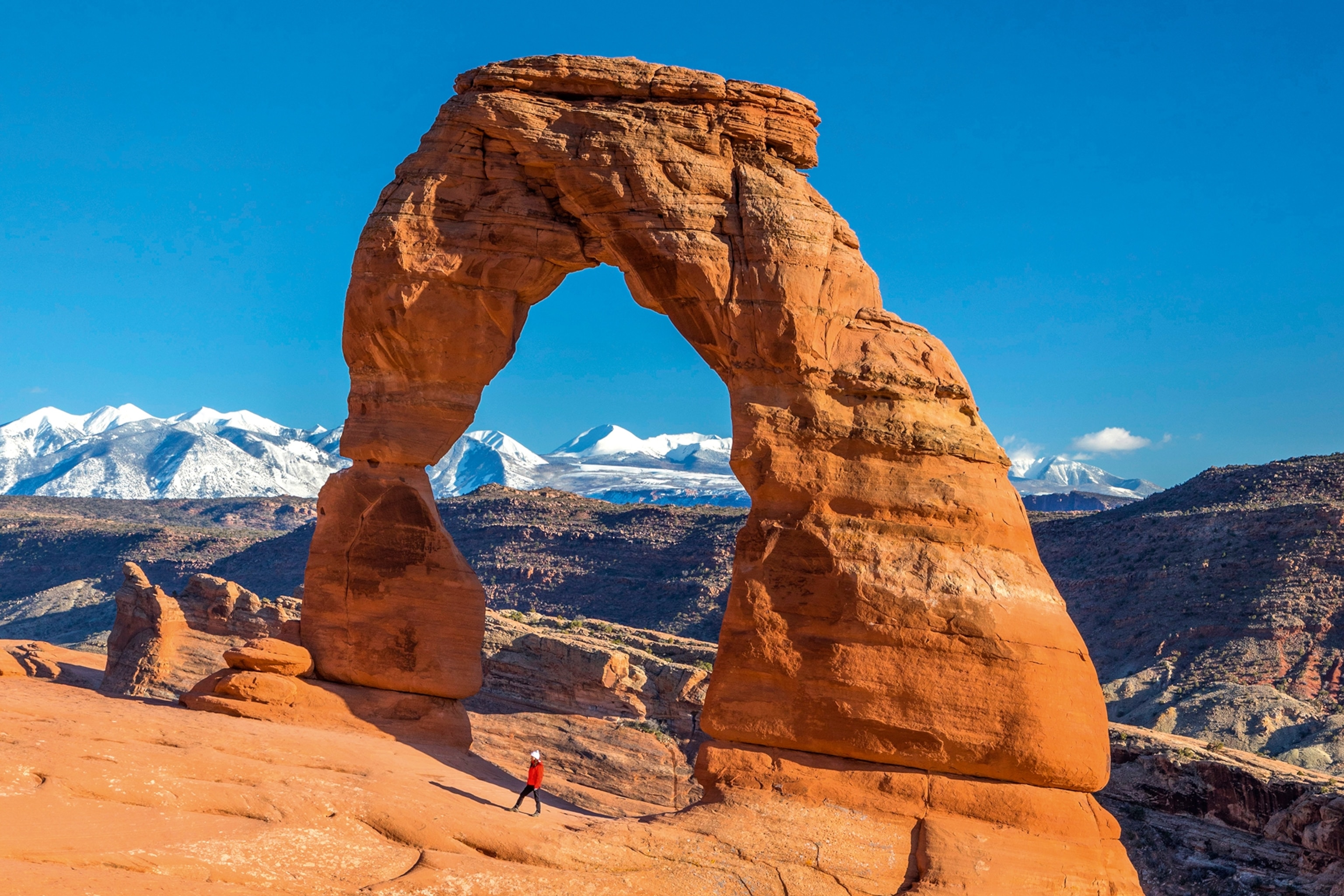 A stone arch monument on the edge of a canyon with a snow-tipped mountain ridge in the far background.
