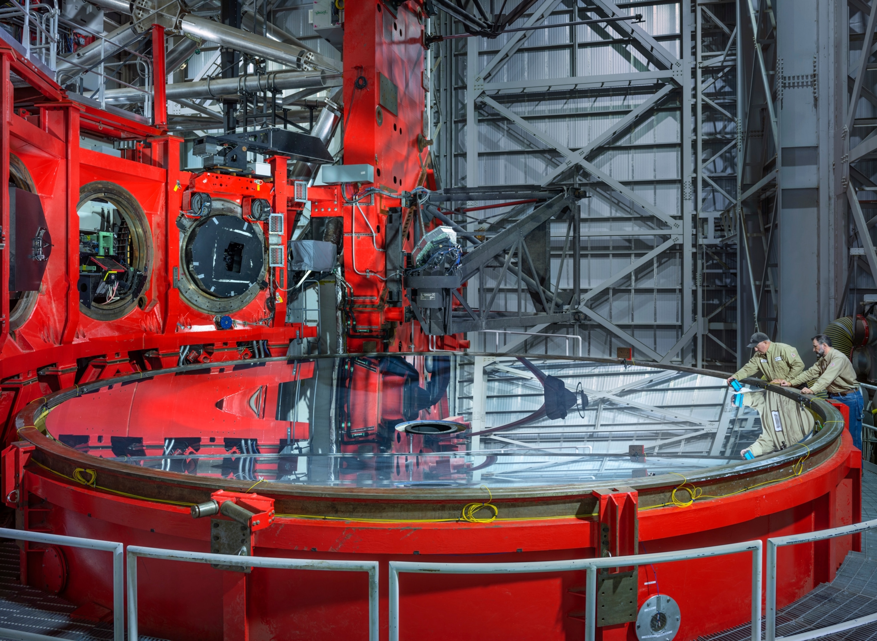 two men in brown shirtfs cleaning the edge of a massive silver disk housed in a complex set of red painted metal structures.