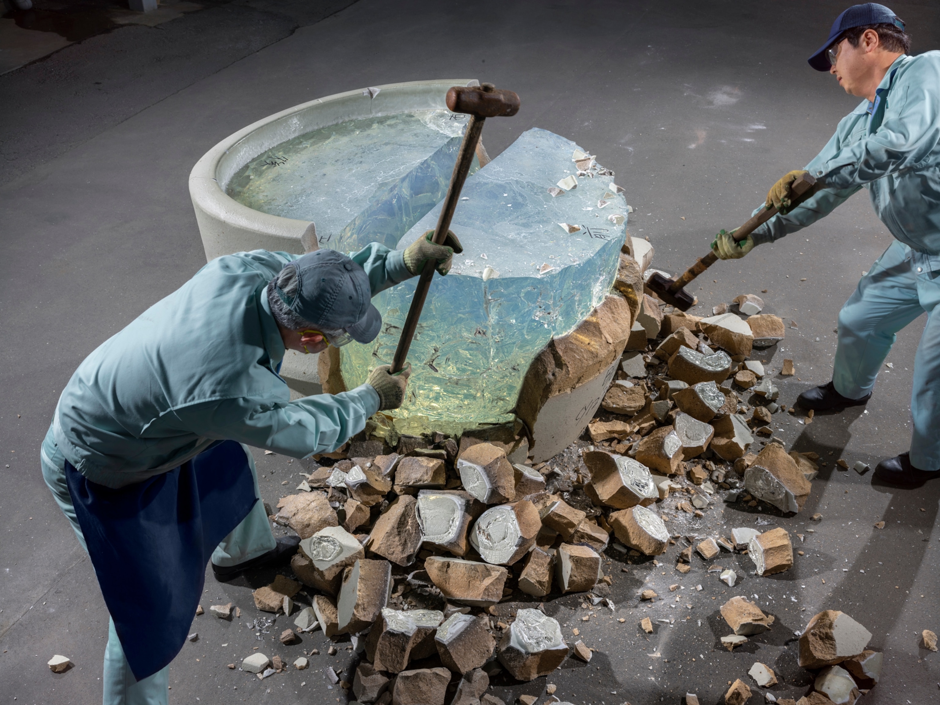 two men in blue suits swing sledgehammers at a massive piece of glass in attempt to remote it from its concrete vessel.