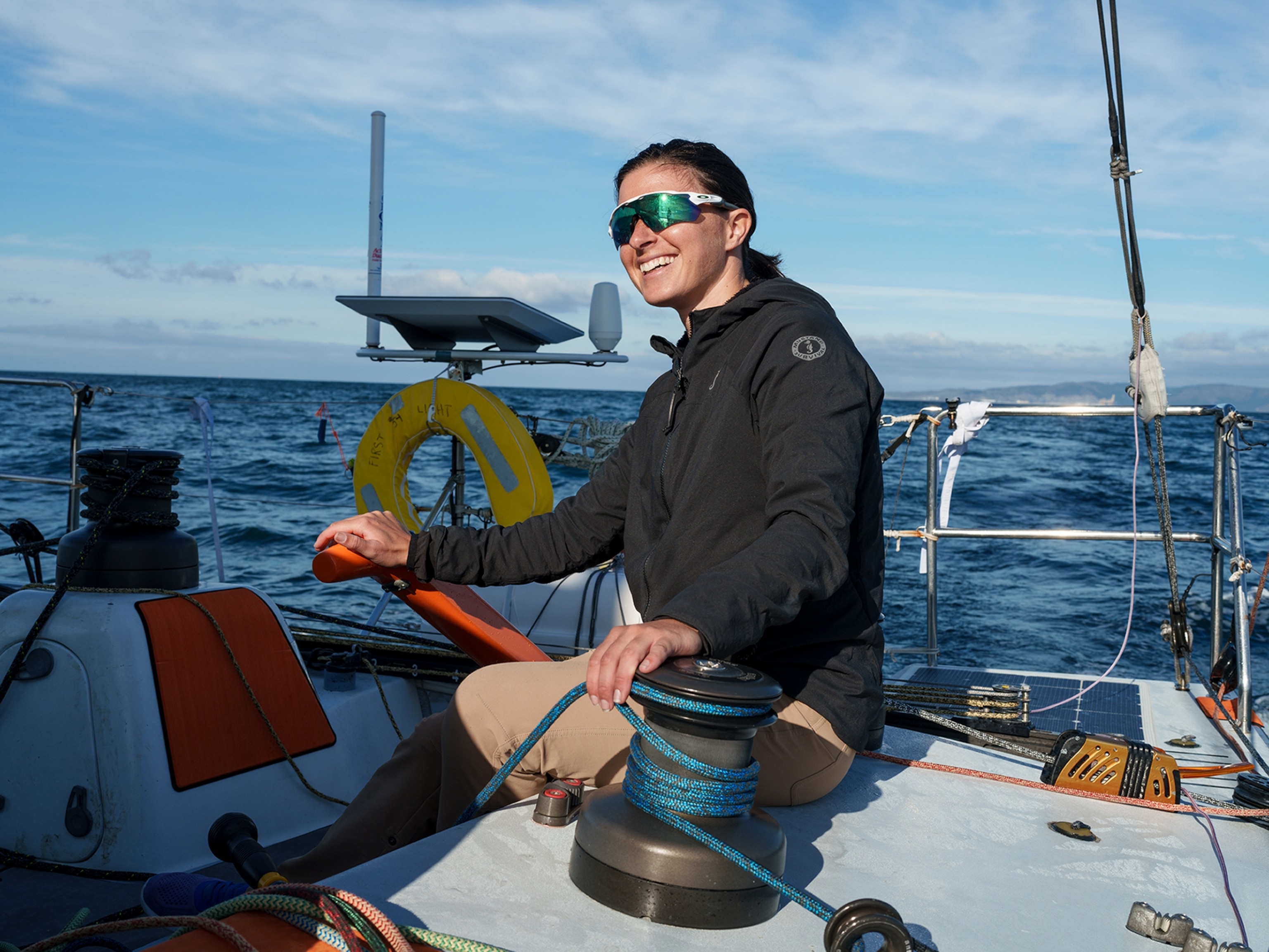 A woman smiles as she steers on a sailboat off the coast. She is wearing sunglasses and the sun is reflected in them. It is a clear and bright day.