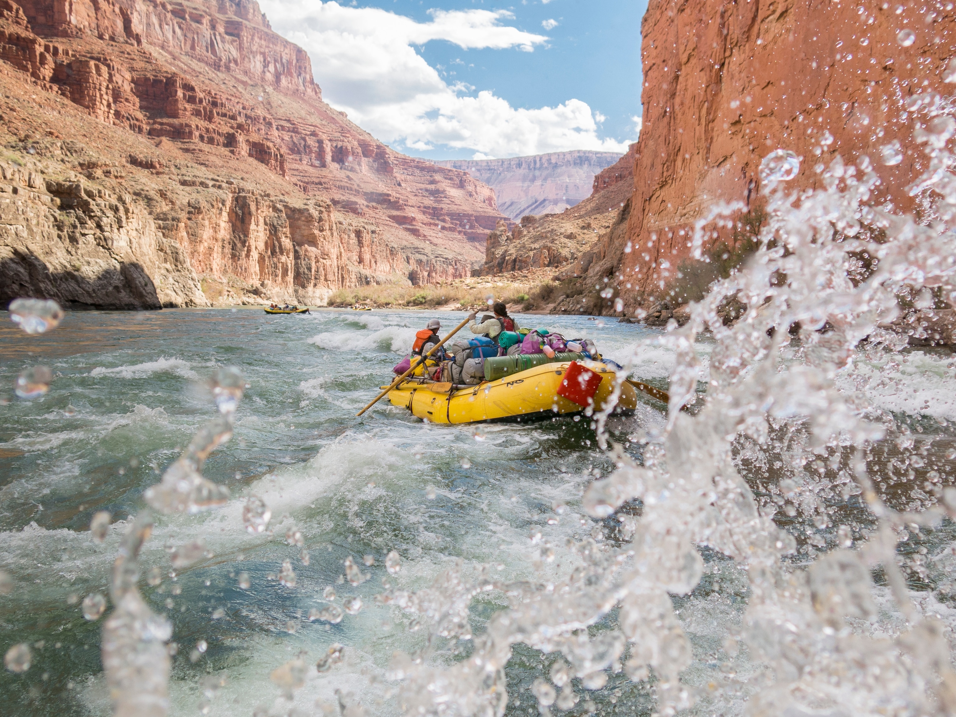 A group of people on a yellow raft, paddling a whitewater raft through rapids on Colorado River in the Grand Canyon.