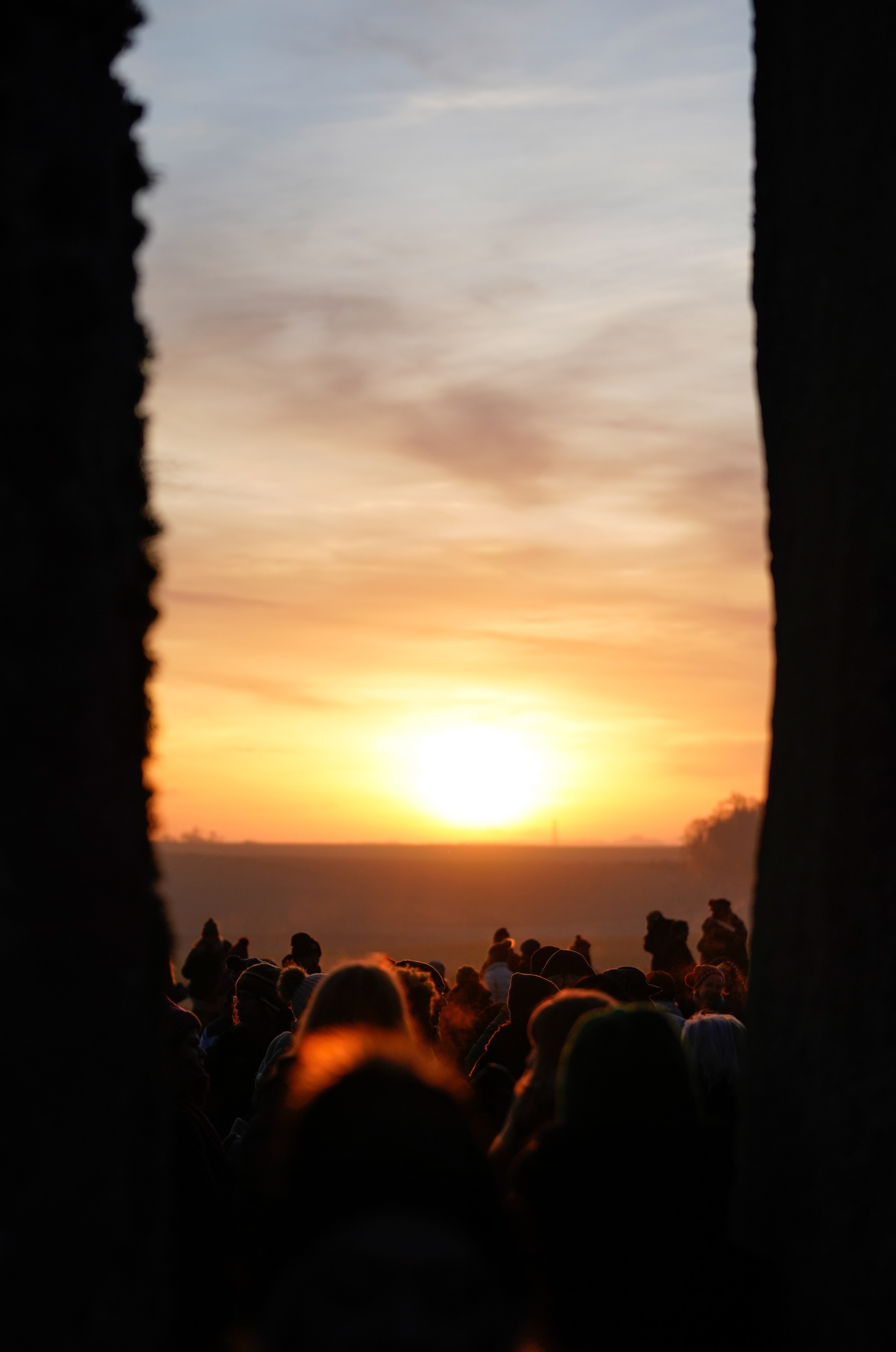 The backs of a group of people watching the sun rise above the horizon are silhouetted between two tall stone megaliths at the left and right of frame.