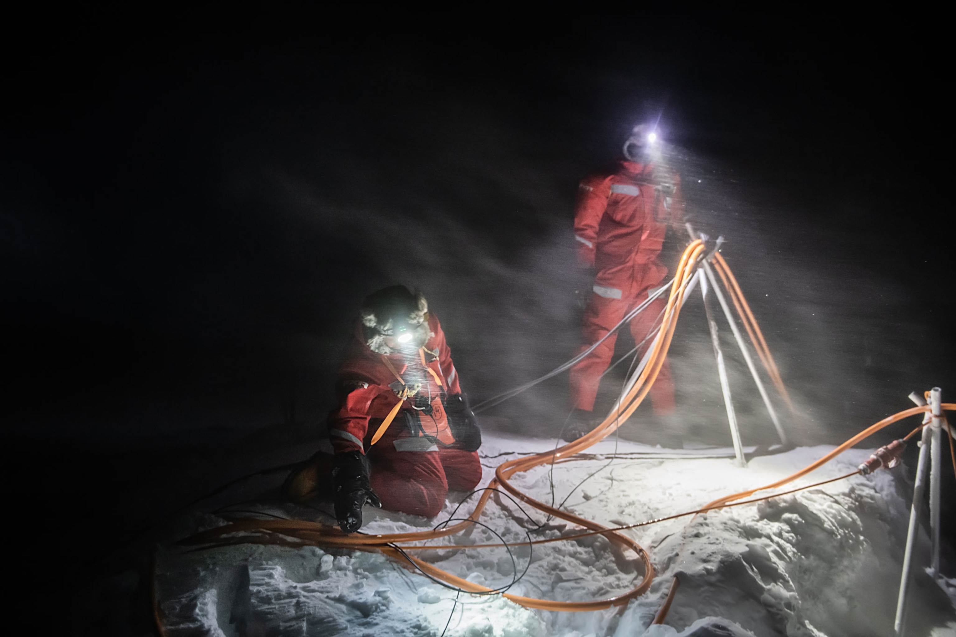 Two people in red snow suits and headlamps work with cables in an arctic snowstorm