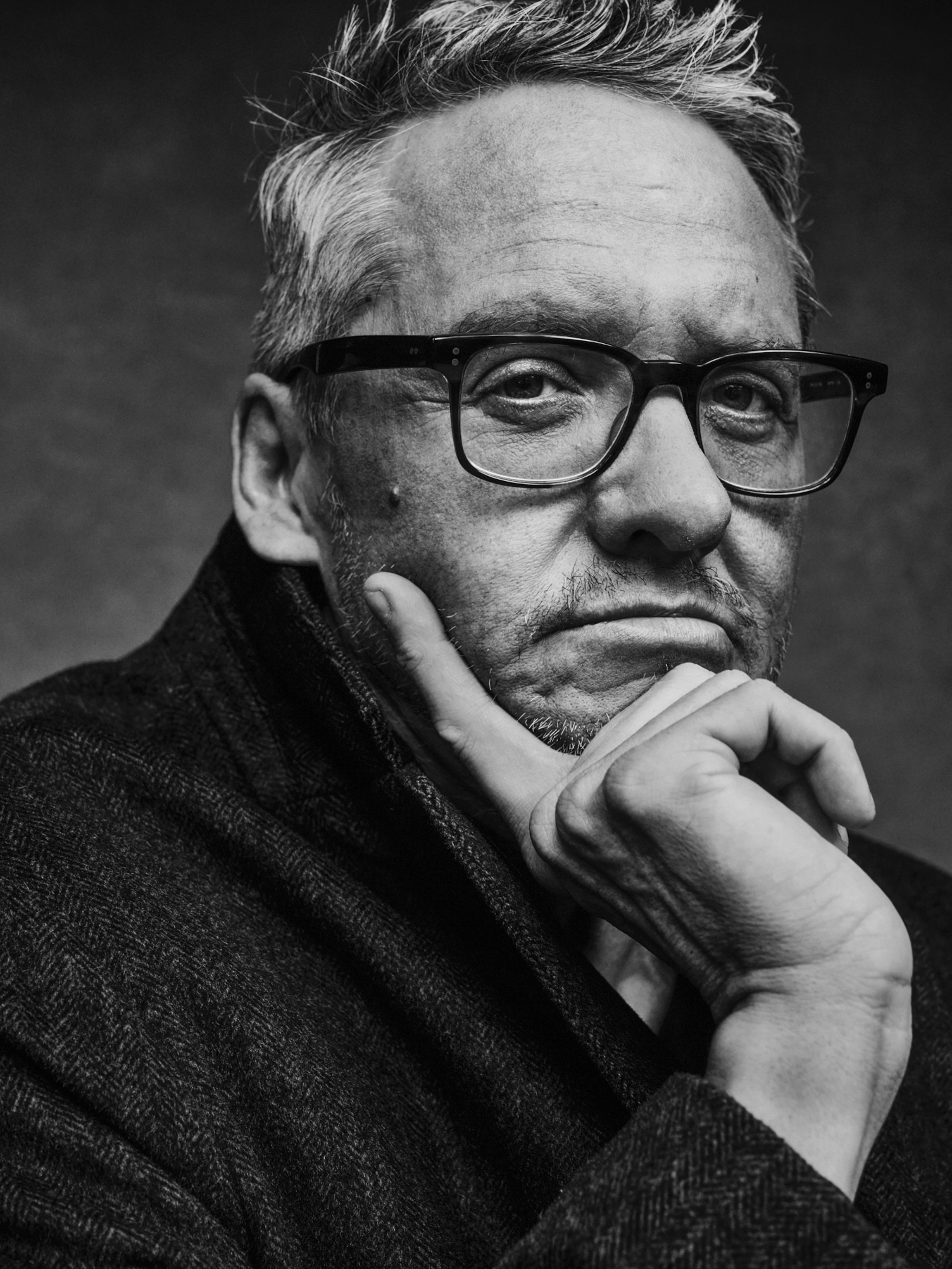 A man sits for a portrait. He looks directly at the camera. He has square glasses and his hand is on his chin. The photo is in black and white.