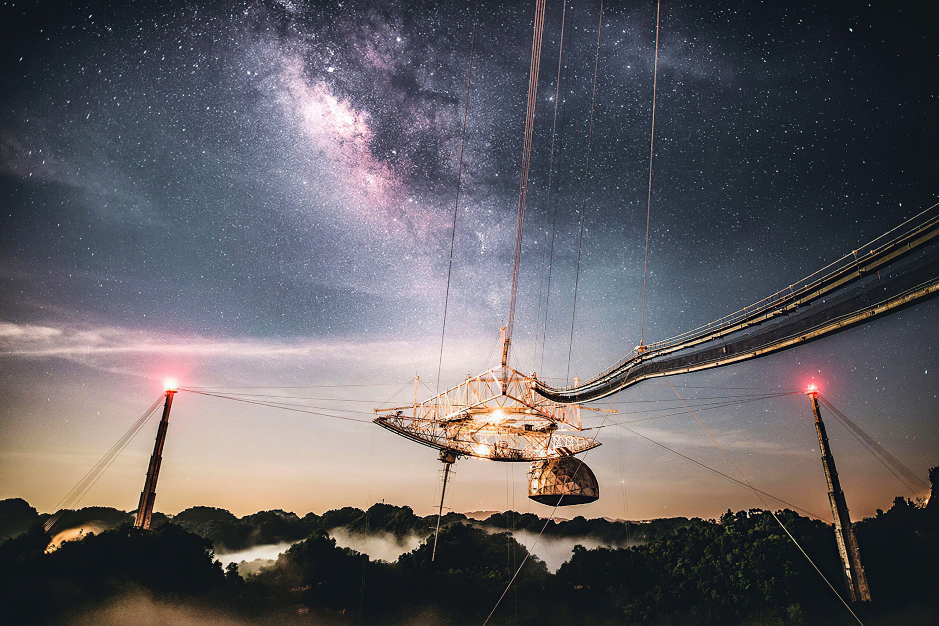 A metallic semi-dome structure suspended from a series of metal scaffolding, suspended between towers aglow with red beacons. Above, the Milky Way fills the night sky.