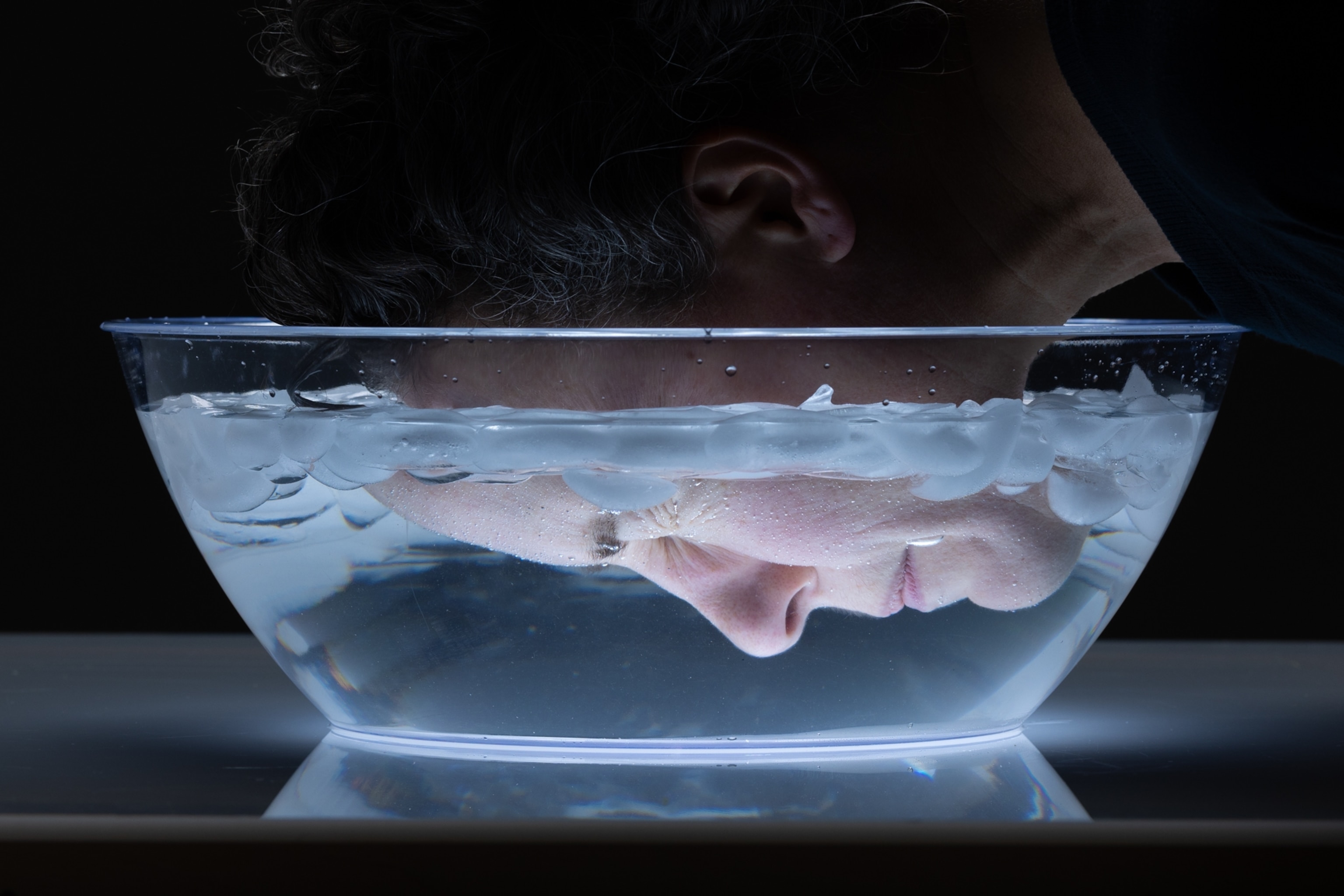 A woman's face, seen from the side, as she submerges her face in a bowl of ice water.