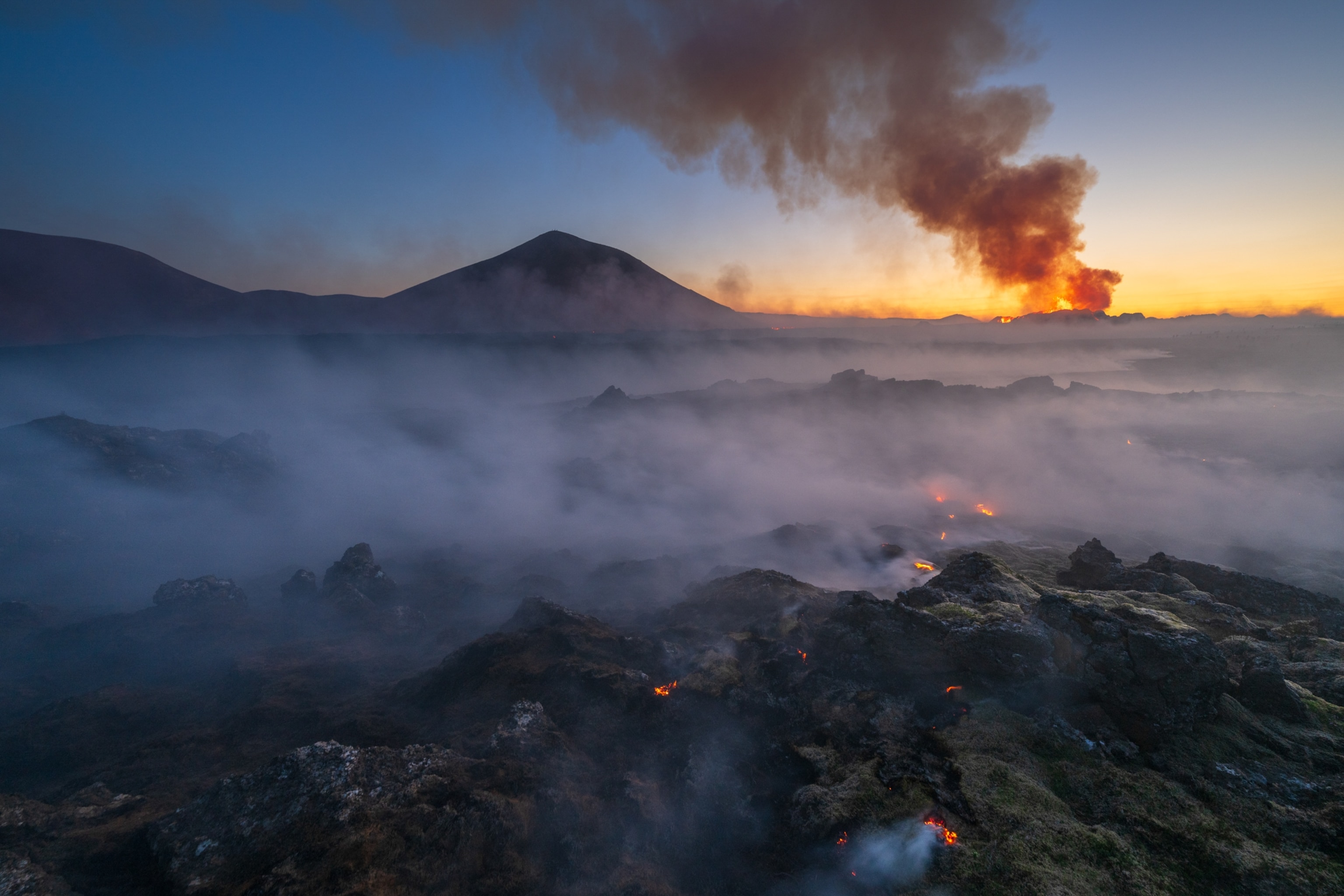 Smoke covers rocky terrain, with an active eruption seen in the distance.