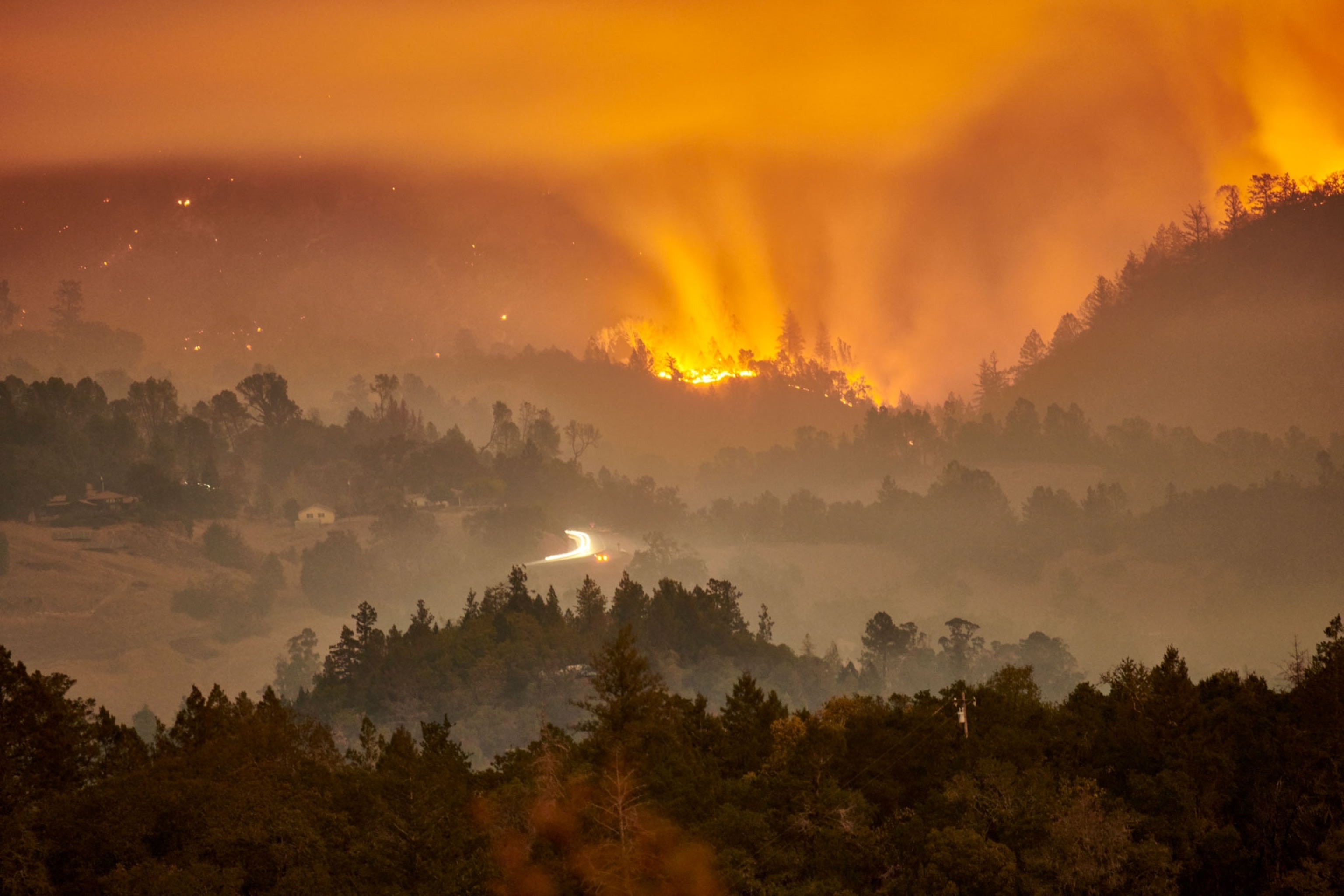 orange and brown tinted smoke fills a valley with the glow of wildfire in the distance