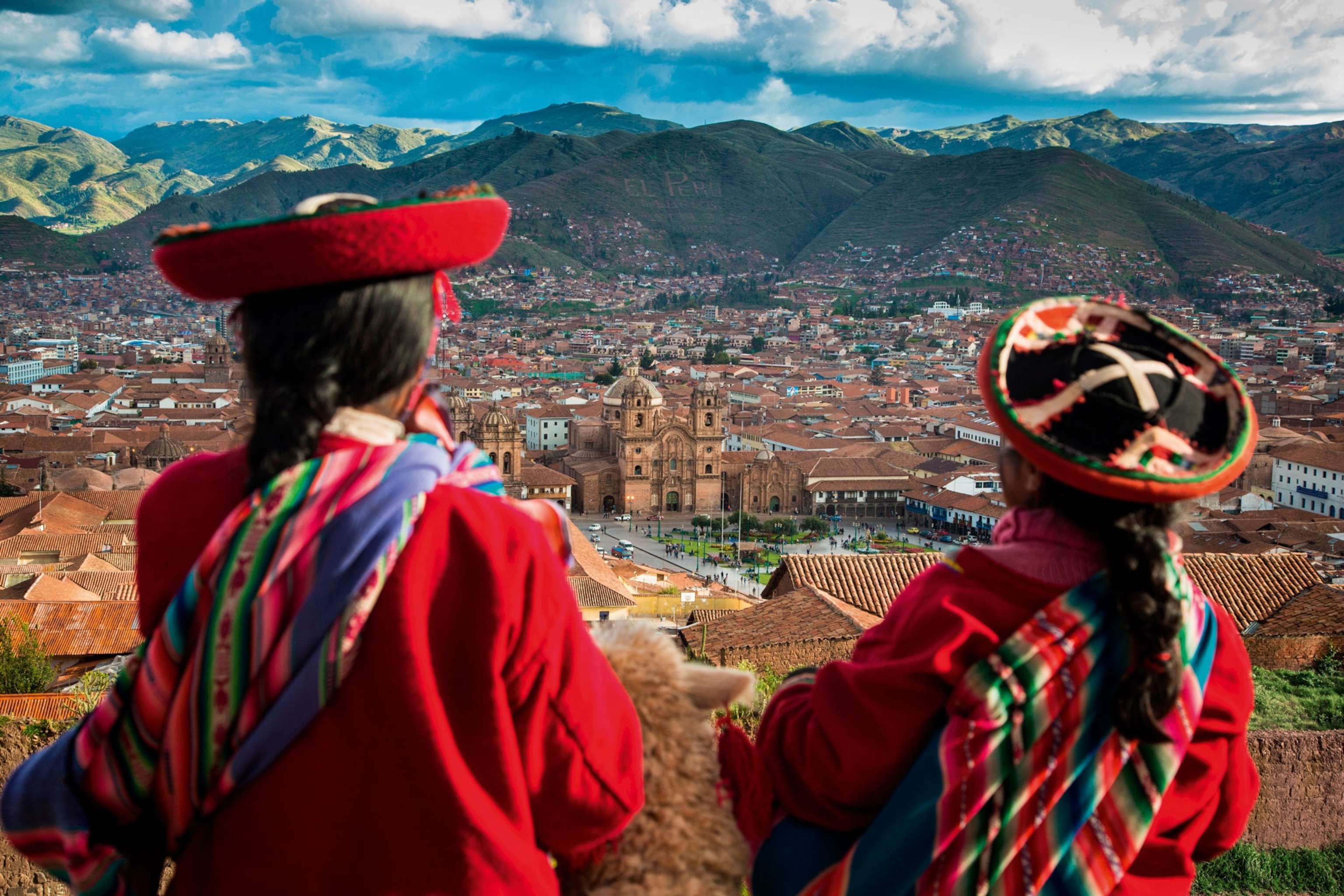 Two people overlooking a historic valley town surrounded by mountains.