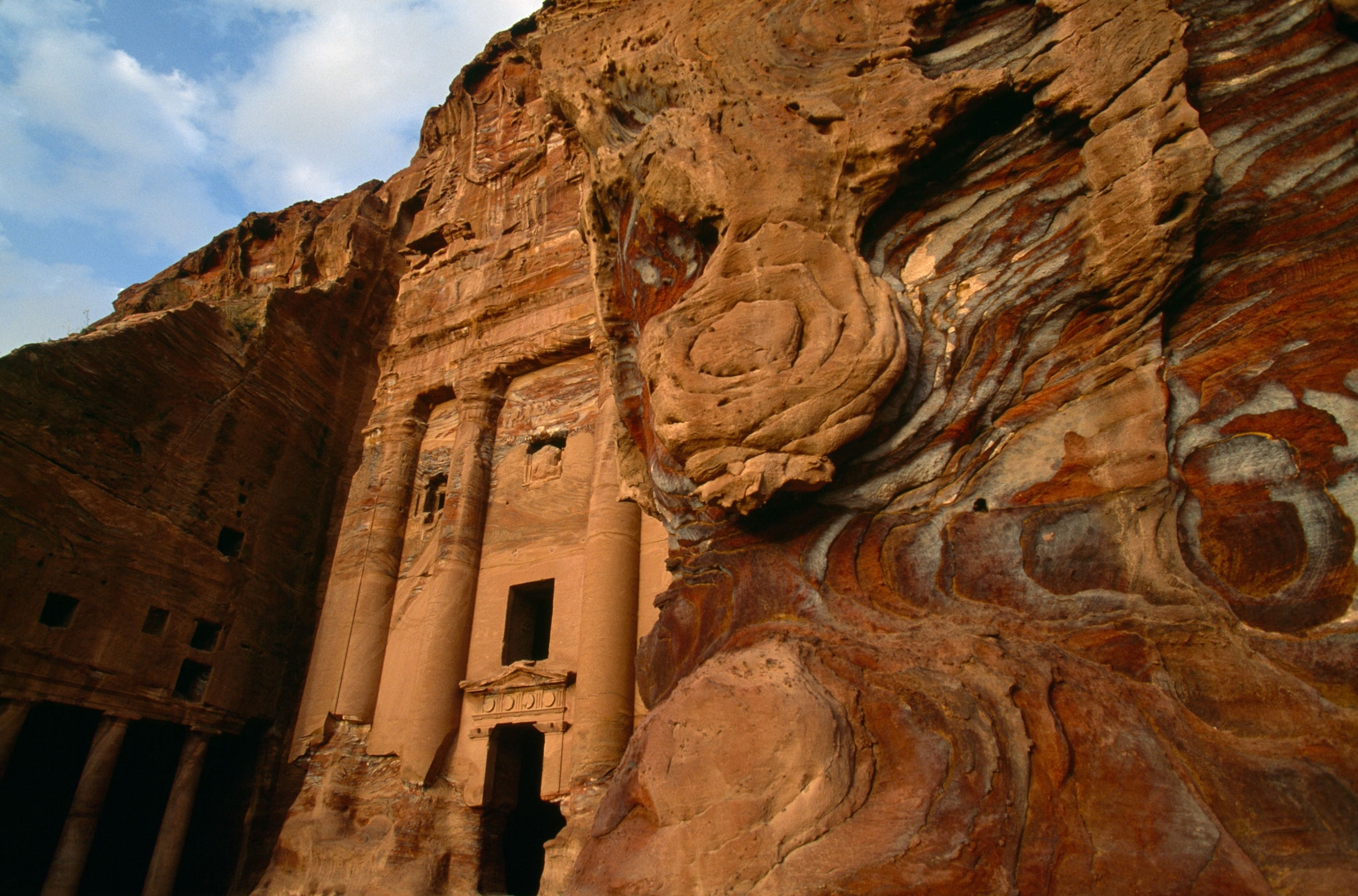 Swirling patterns of reds and browns appear in eroded sandstone in the foreground, while part of a facade carved into the rock wall is visible in the background