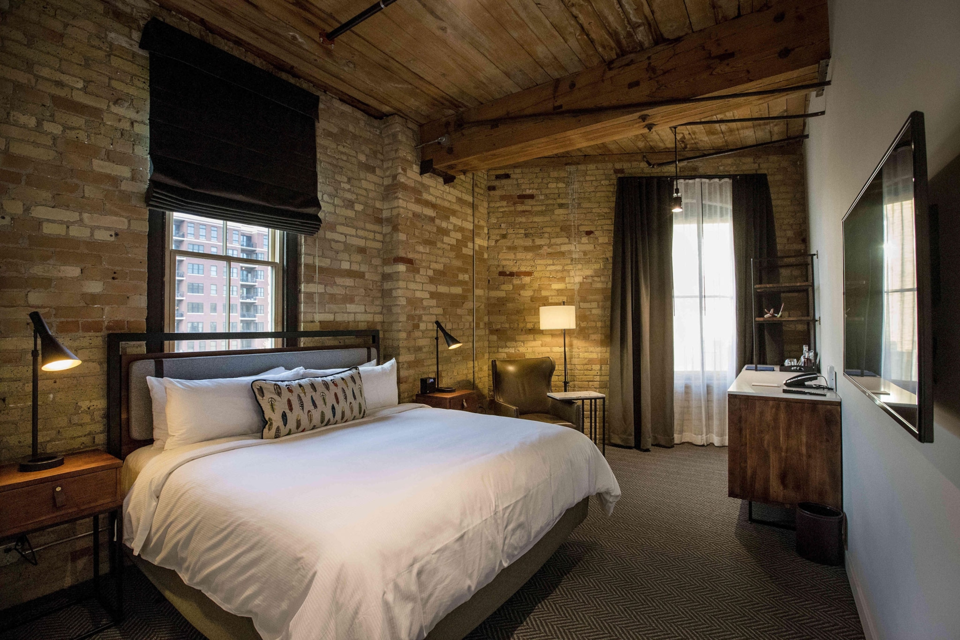 A corner hotel room with grey brick walls, a large bed with white bedding, side lamps, and a desk.