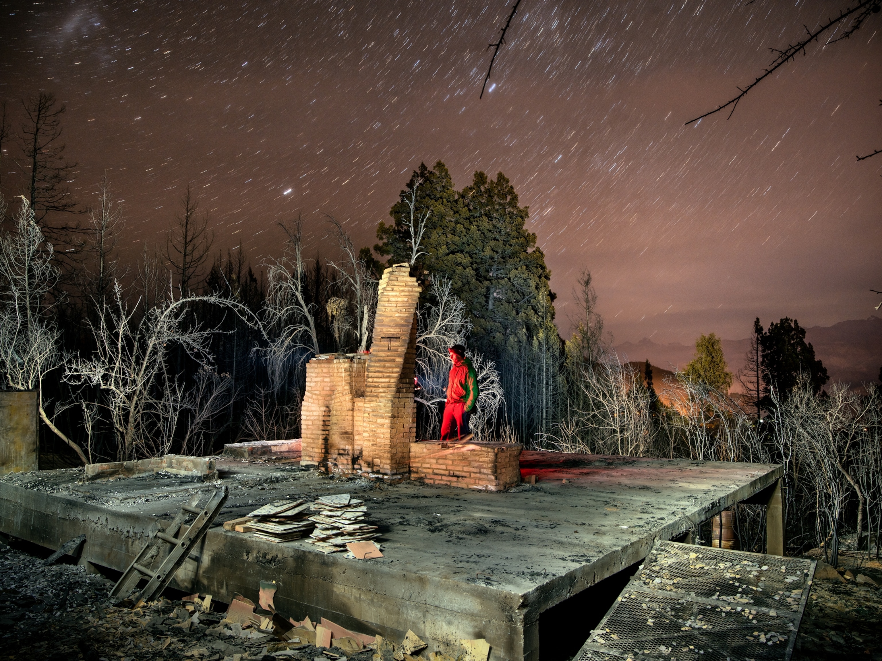 a man stands in front of a fire place, all that remains of his home, illuminated in the night by a red lantern