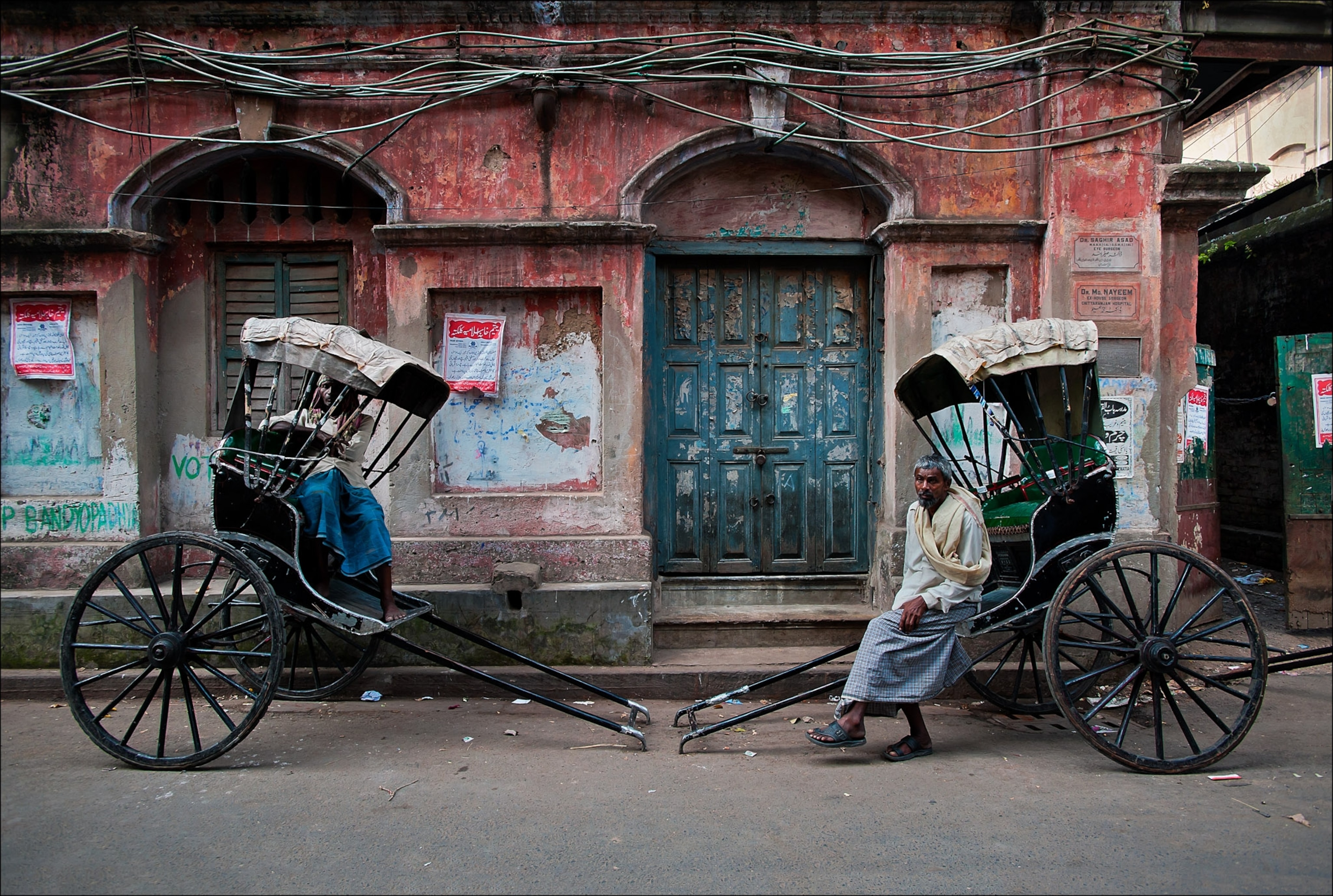 Two men and their rickshaws one sits on the interior obstructed and another sits at edge of his kart.