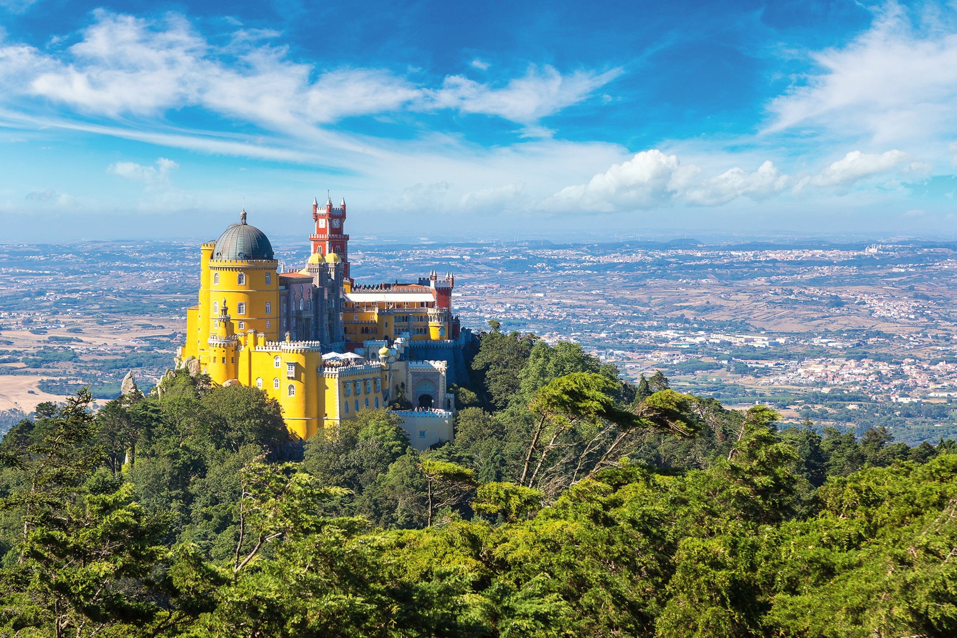 Yellow castle stands surrounded by trees with a backdrop of countryside and towns below