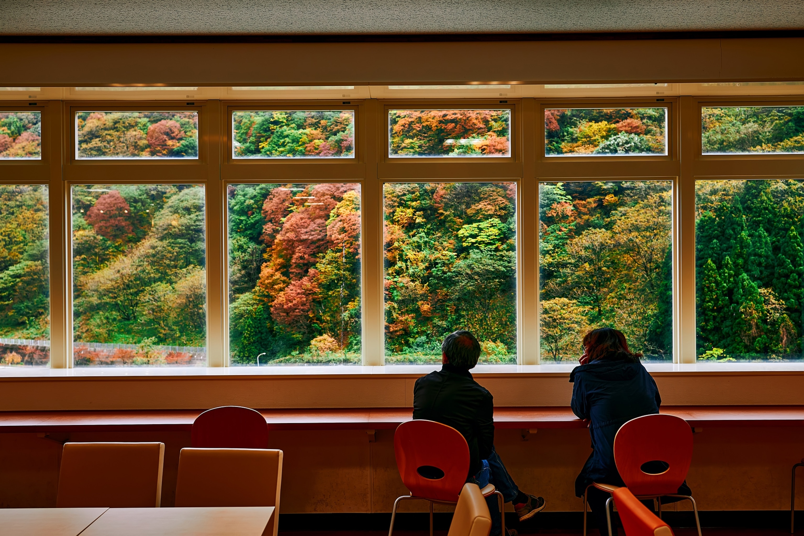 A photograph of two people seated in Unazuki station looking at view of Kurobe Gorge which features beautiful fall foliage