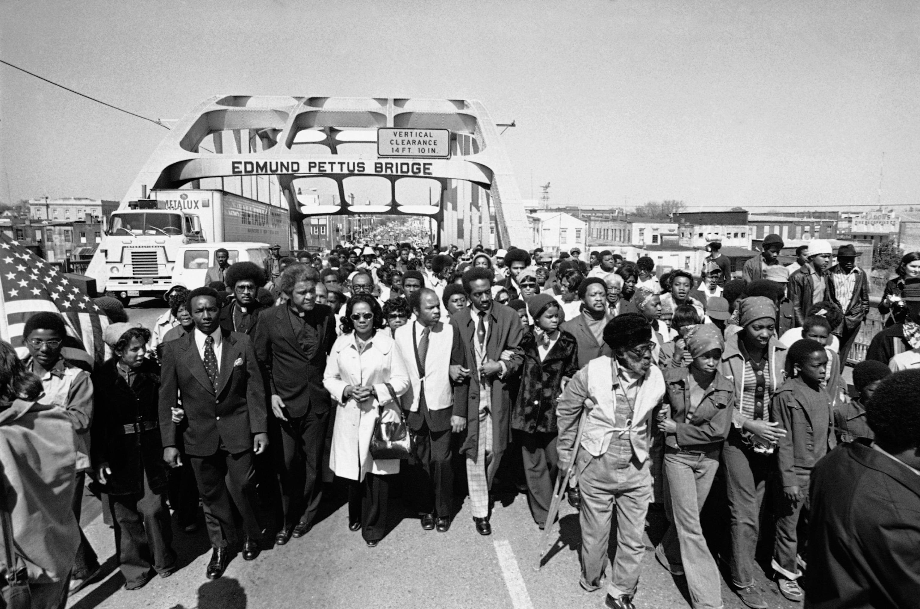 A black and white photograph of of a group of people with dark skin linked arm and arm walking across a bridge the crowd continues into the distance filling the walkway.