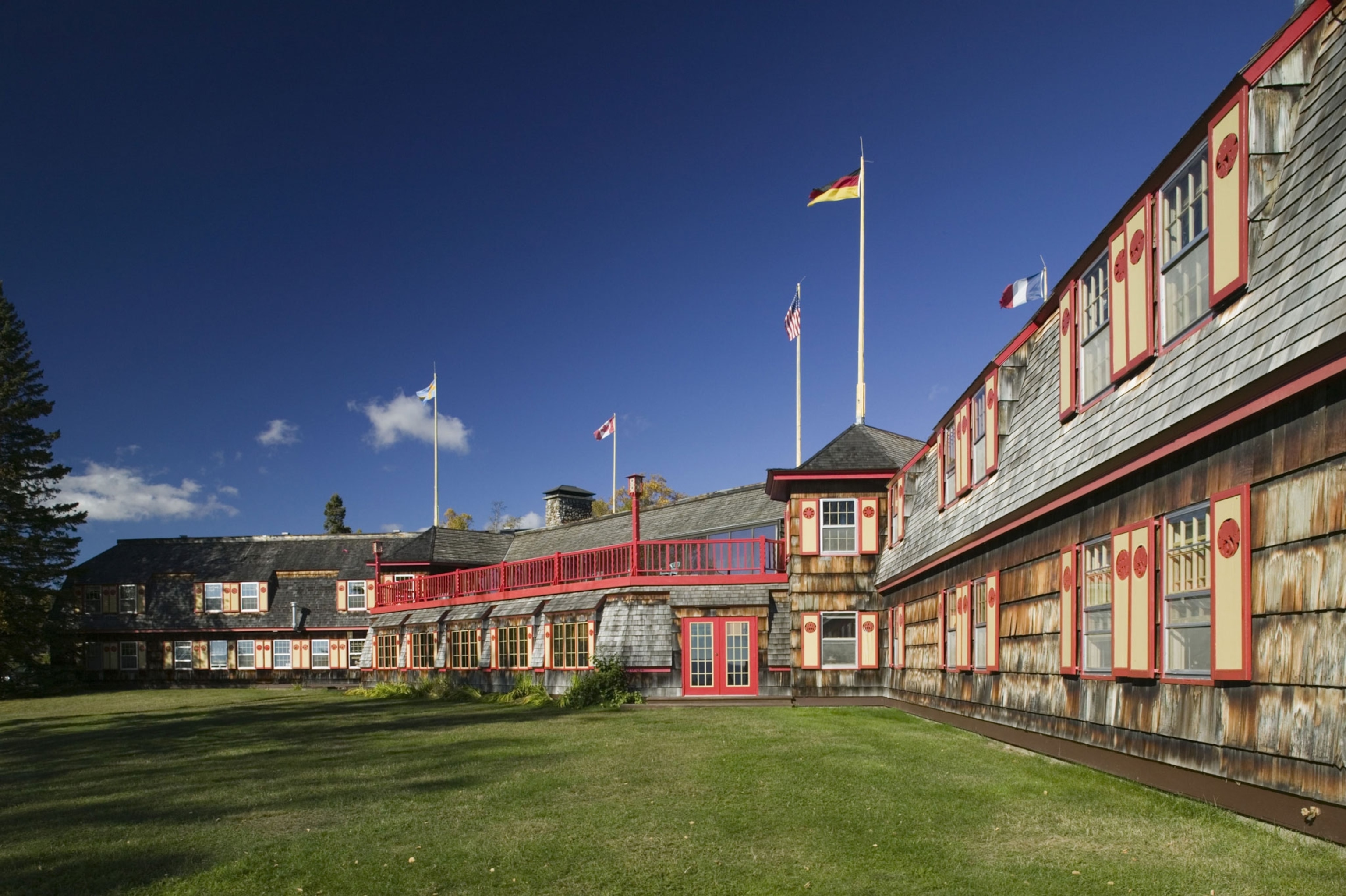 A two story hotel with wood siding, red trimmed yellow shutters, that extends from the foreground to the background. The hotel is surround with green grass and a pine tree off to the side.