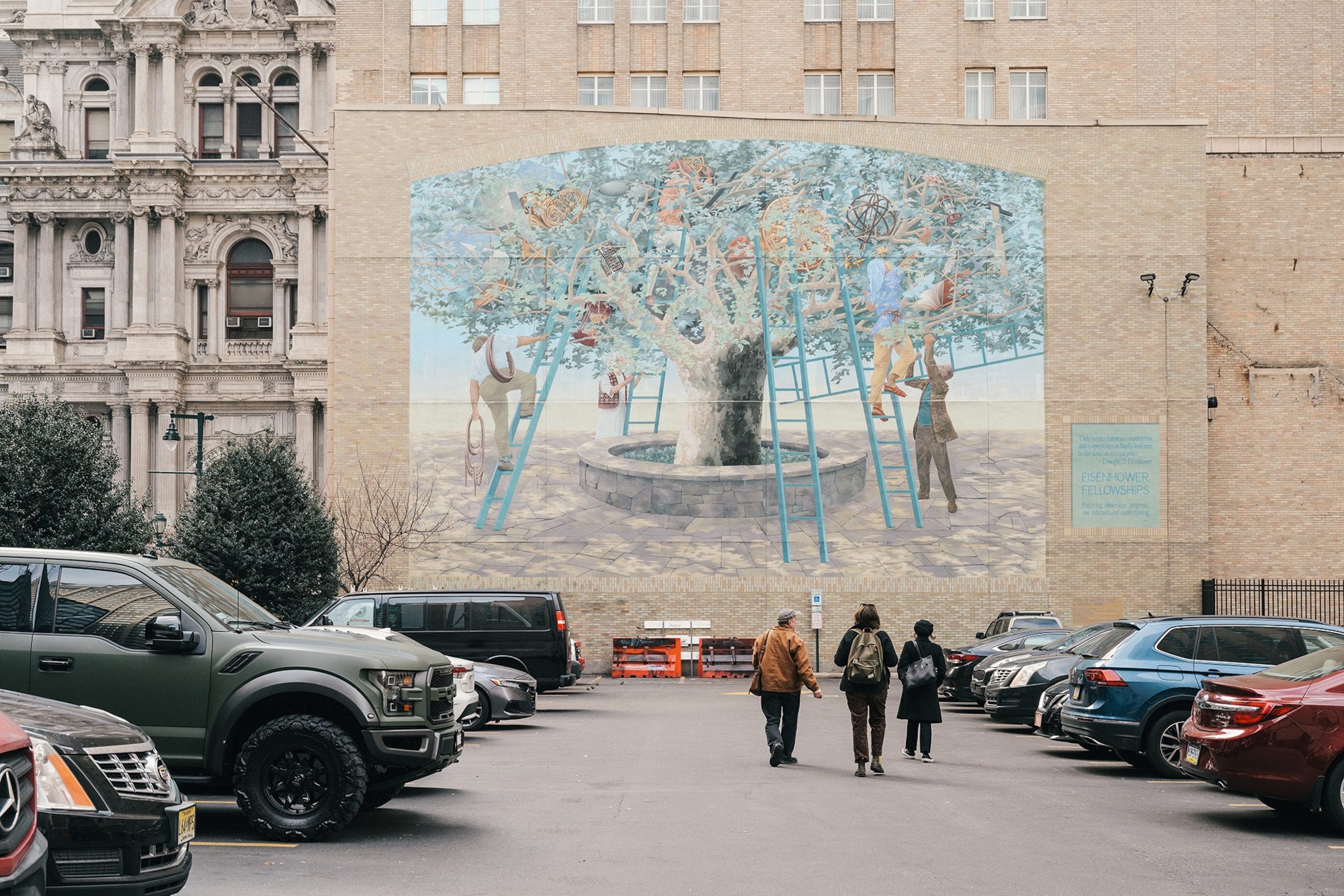 A tree mural on the side of a brick building is seen from a car park in Philadelphia. Three people are walking towards the mural with parked cars on either side of them.