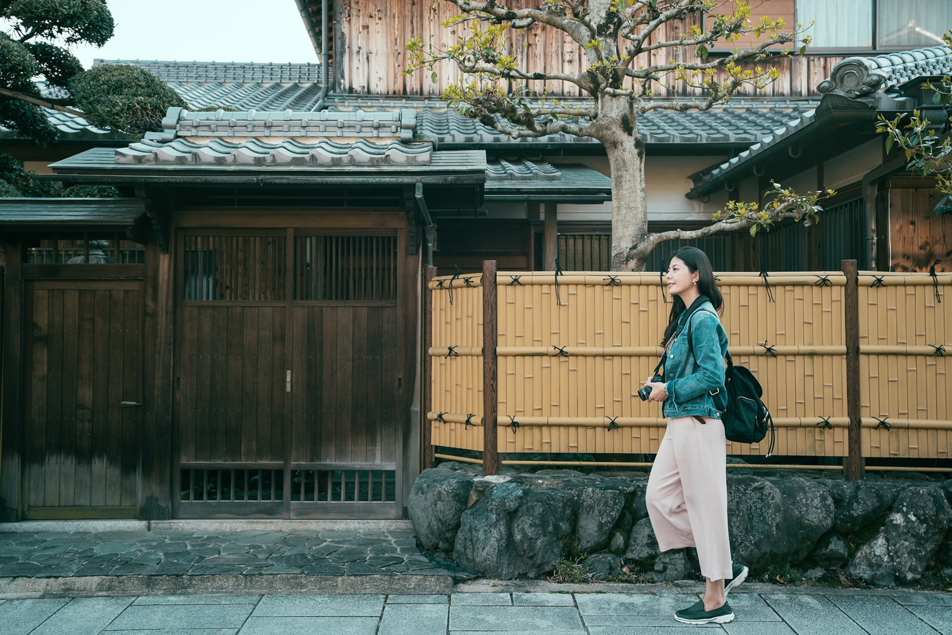 Travel photographer walking along the bamboo walls of a Japanese-style wooden house with trees and a garden inside.