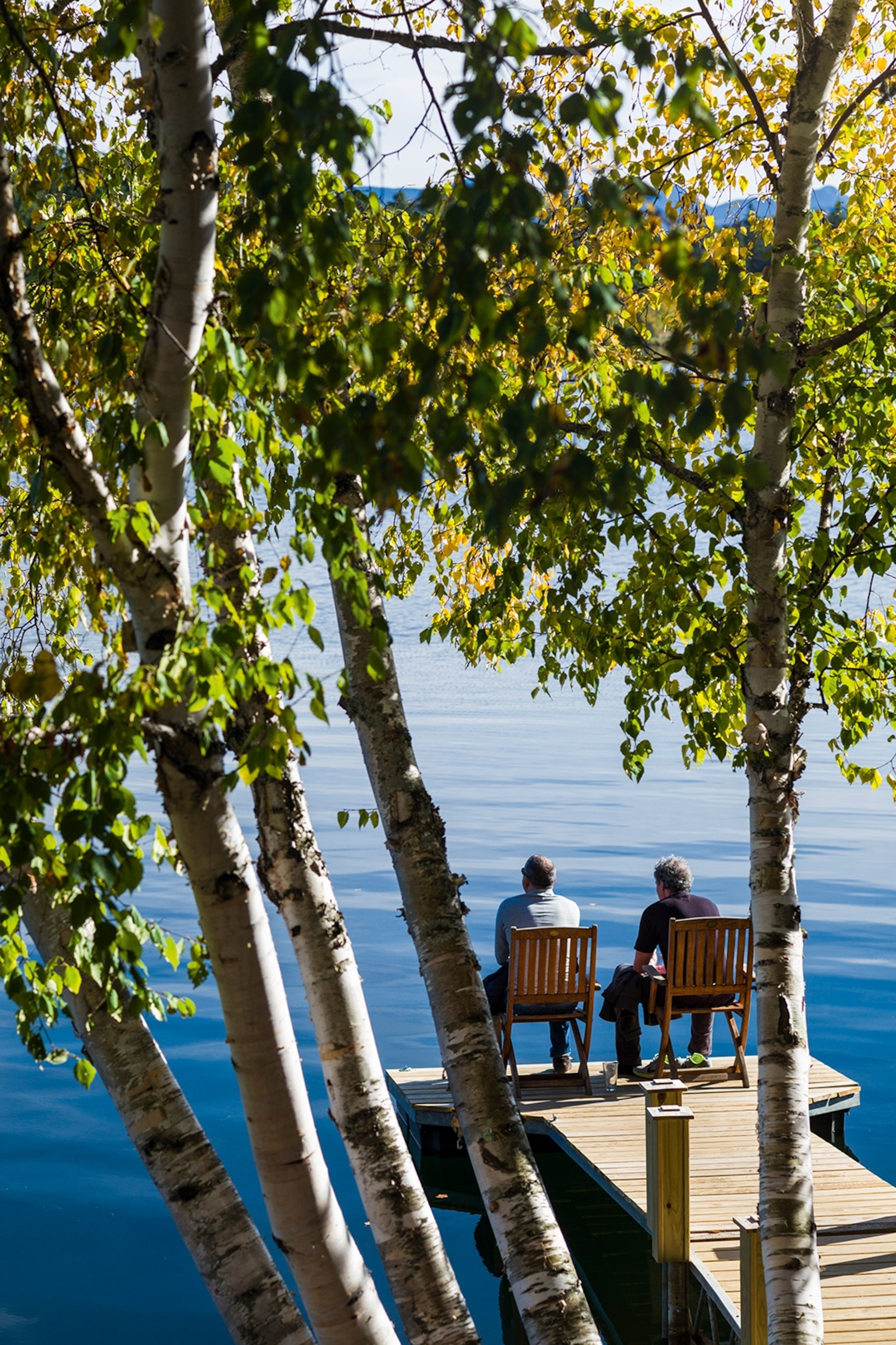 Two men sitting on chairs at the end of a wooden pier, looking out to the lake.
