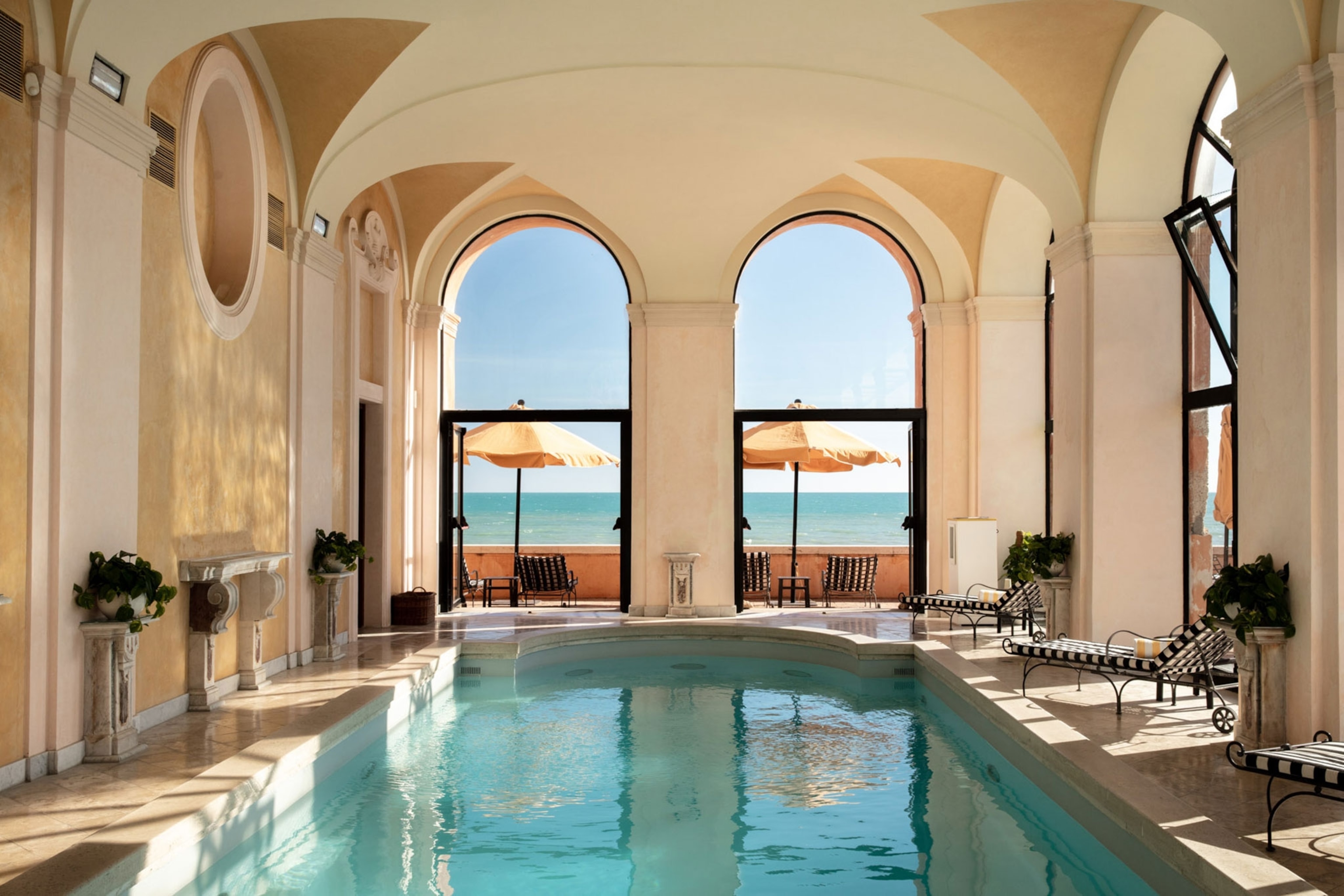 An indoor pool with a view of the ocean outside of the arched windows.