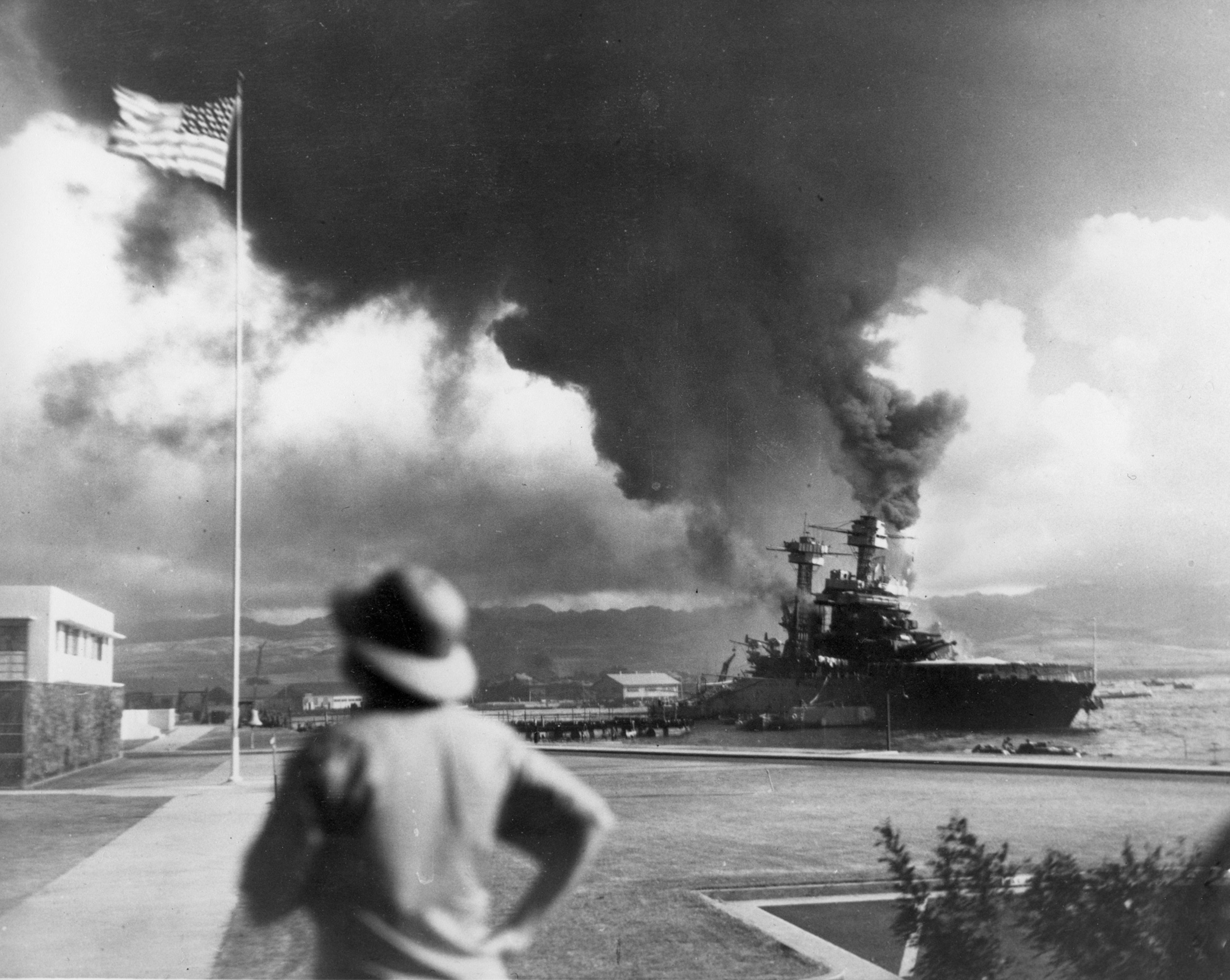 Black smoke billowing from an American ship as a soldier looks on from the foreground.