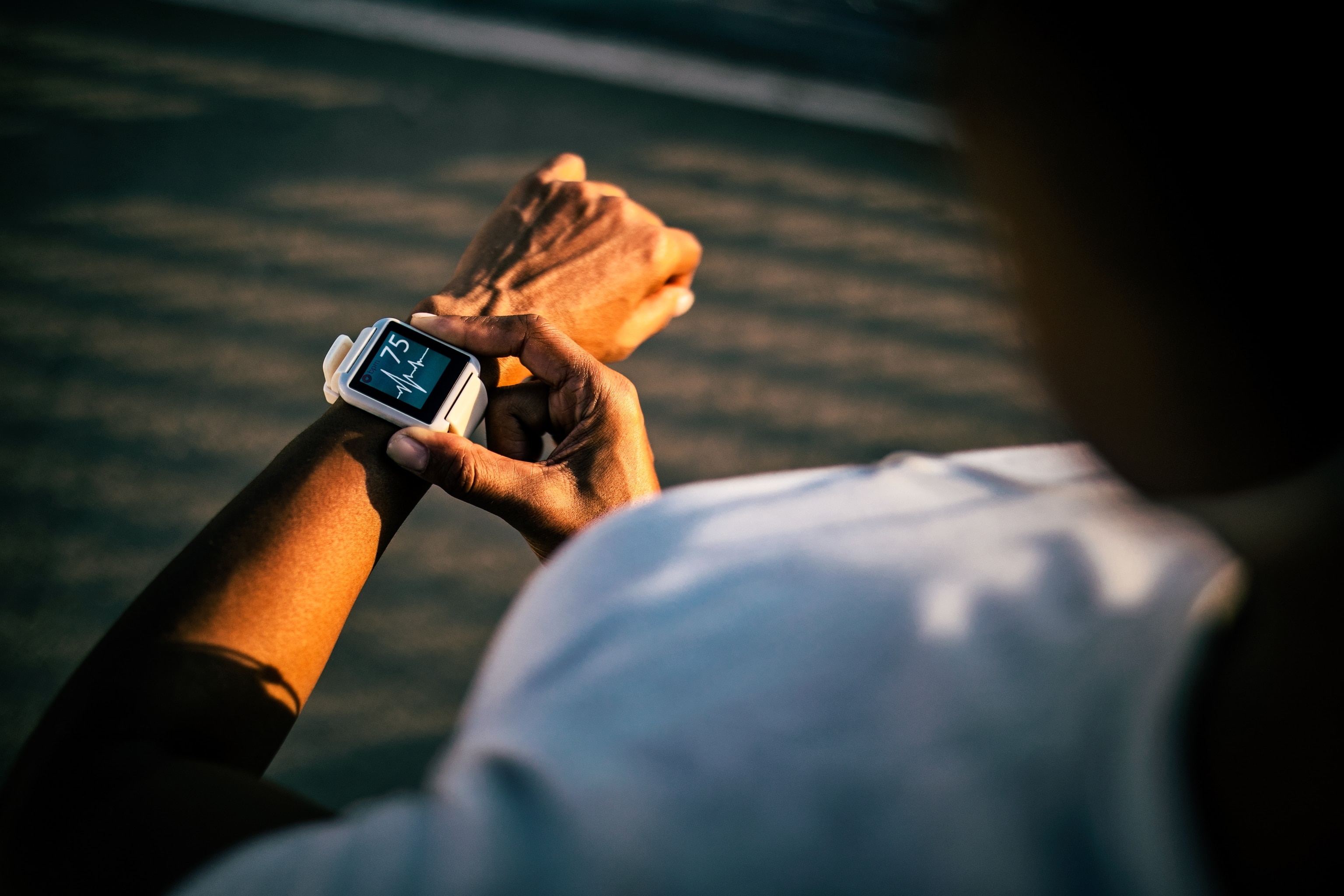 An over-shoulder view of a woman checking her heart-rate using a smart watch, which displays a EKG signal and a heart rate of 75bpm. The woman is standing in dappled sunlight, which primarily illuminates her forearm and smartwatch.
