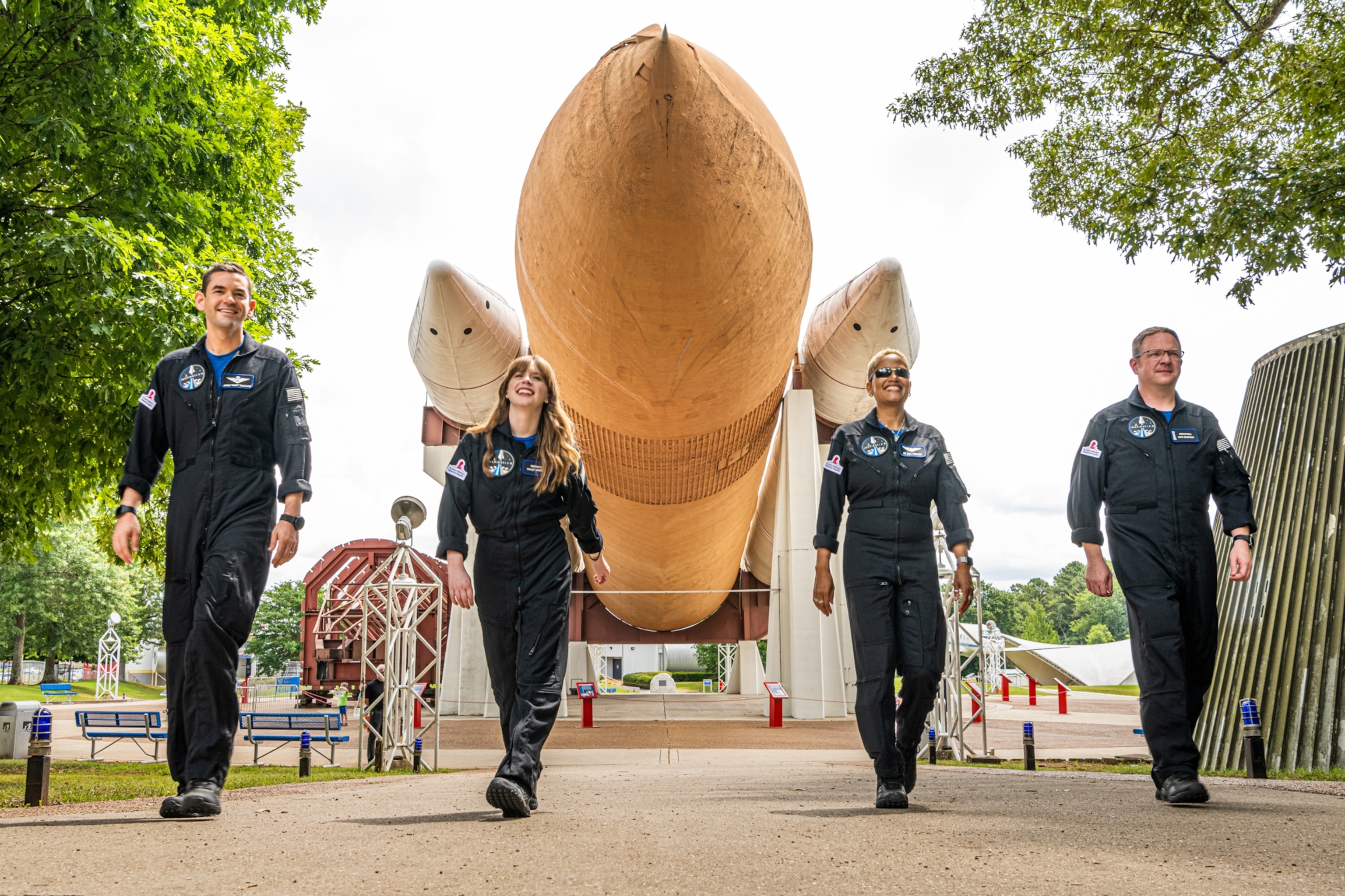 The four crew members of Inspiration4 walking towards the camera. Behind them, a large rocket is displayed, its nose cone pointed towards the camera.