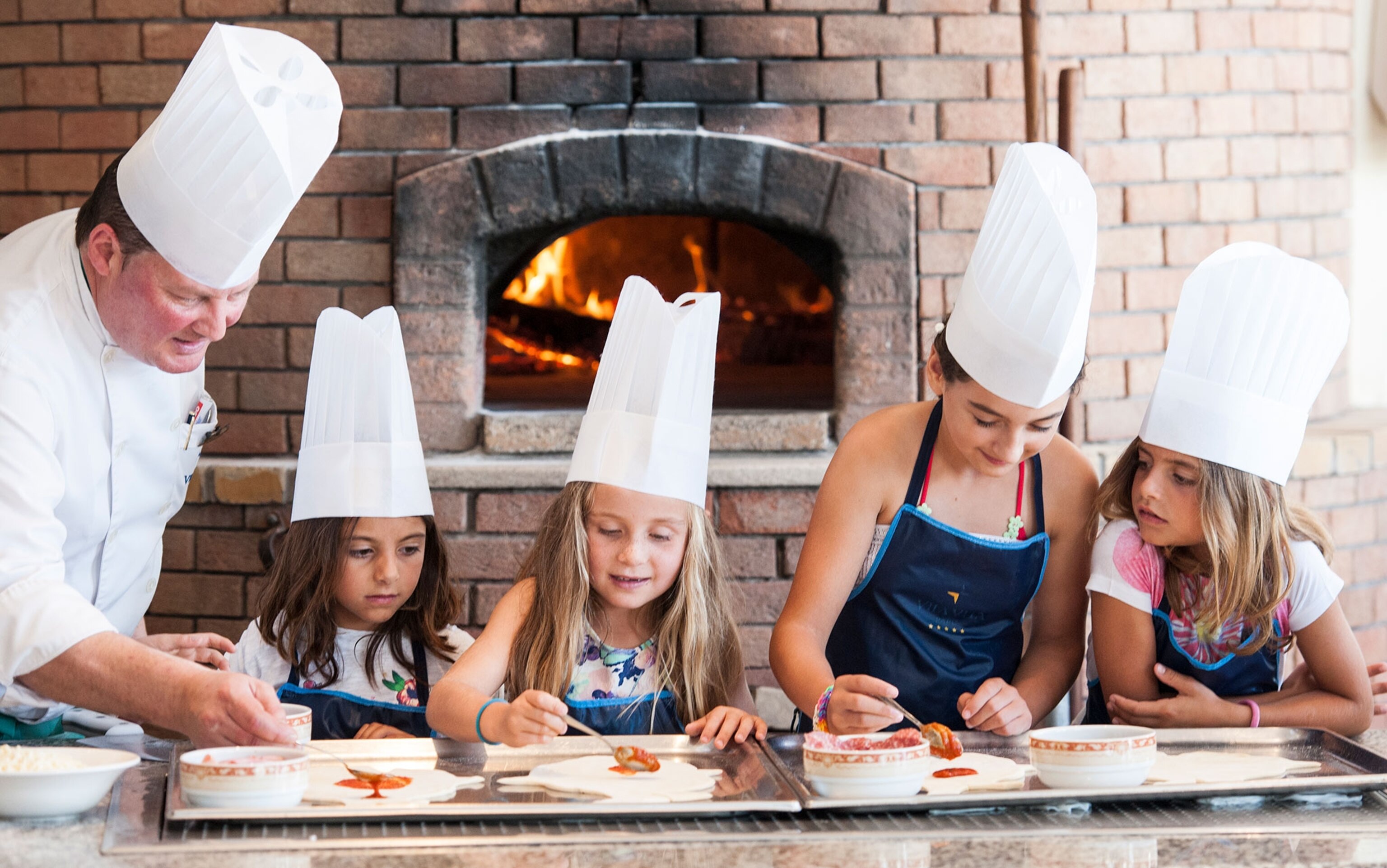 A group of kids and an older man in chef hats spread sauce on pizza dough.