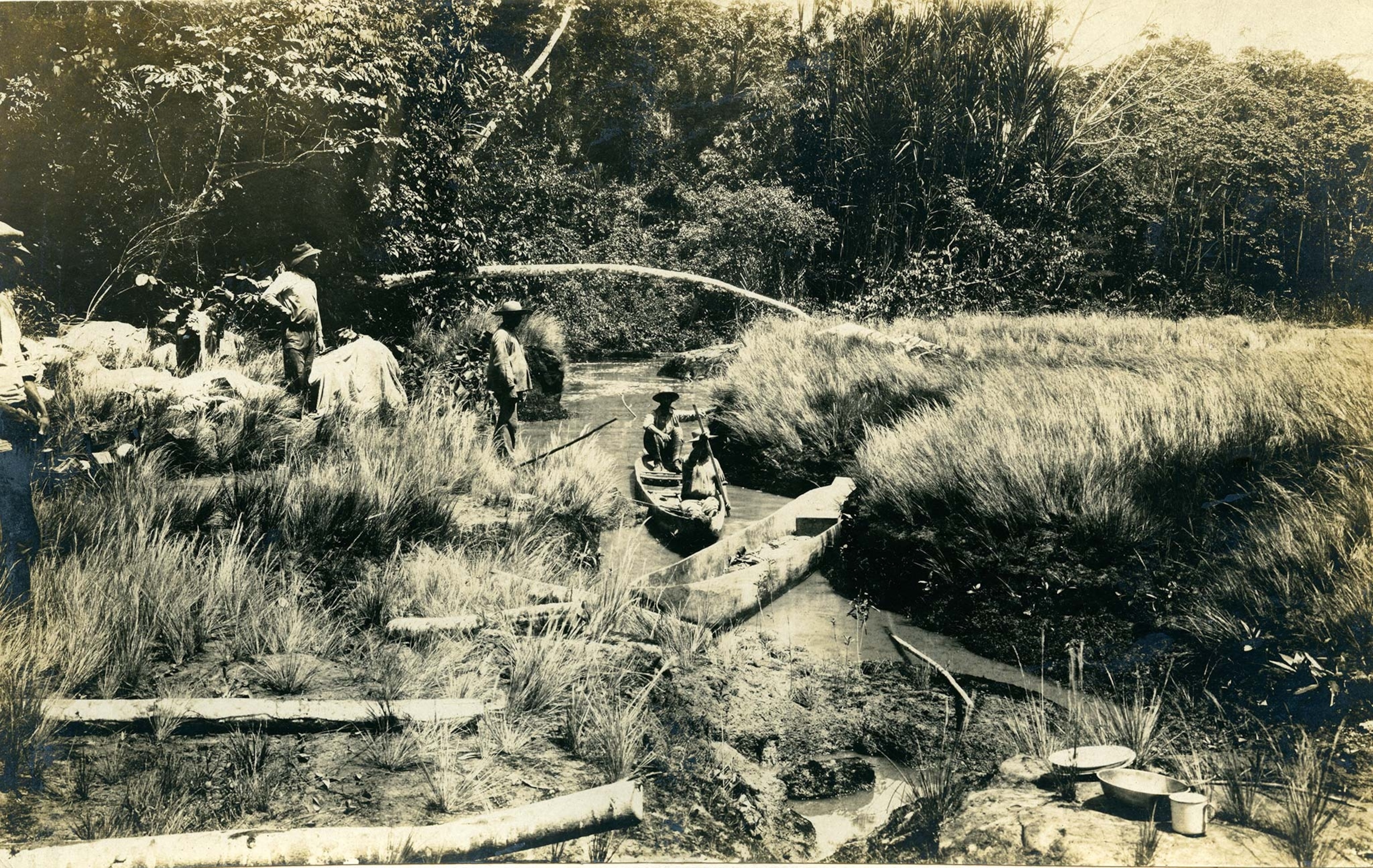Men stand on the shore of a stream and two others sit in a canoe in the water