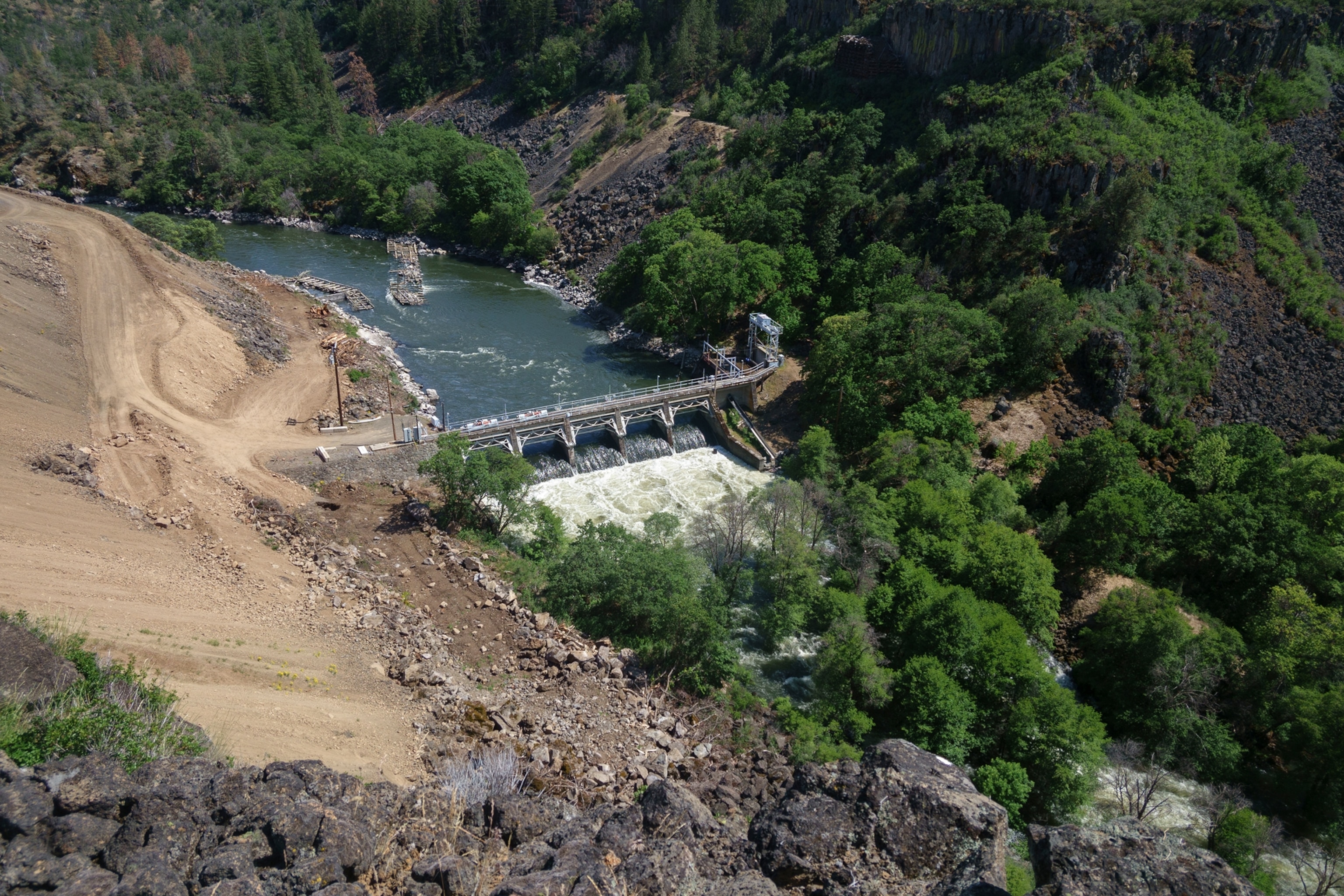 A birds eye view image of a dam in a river. One side is heavily forested, the other is more barren with dirt and rock.