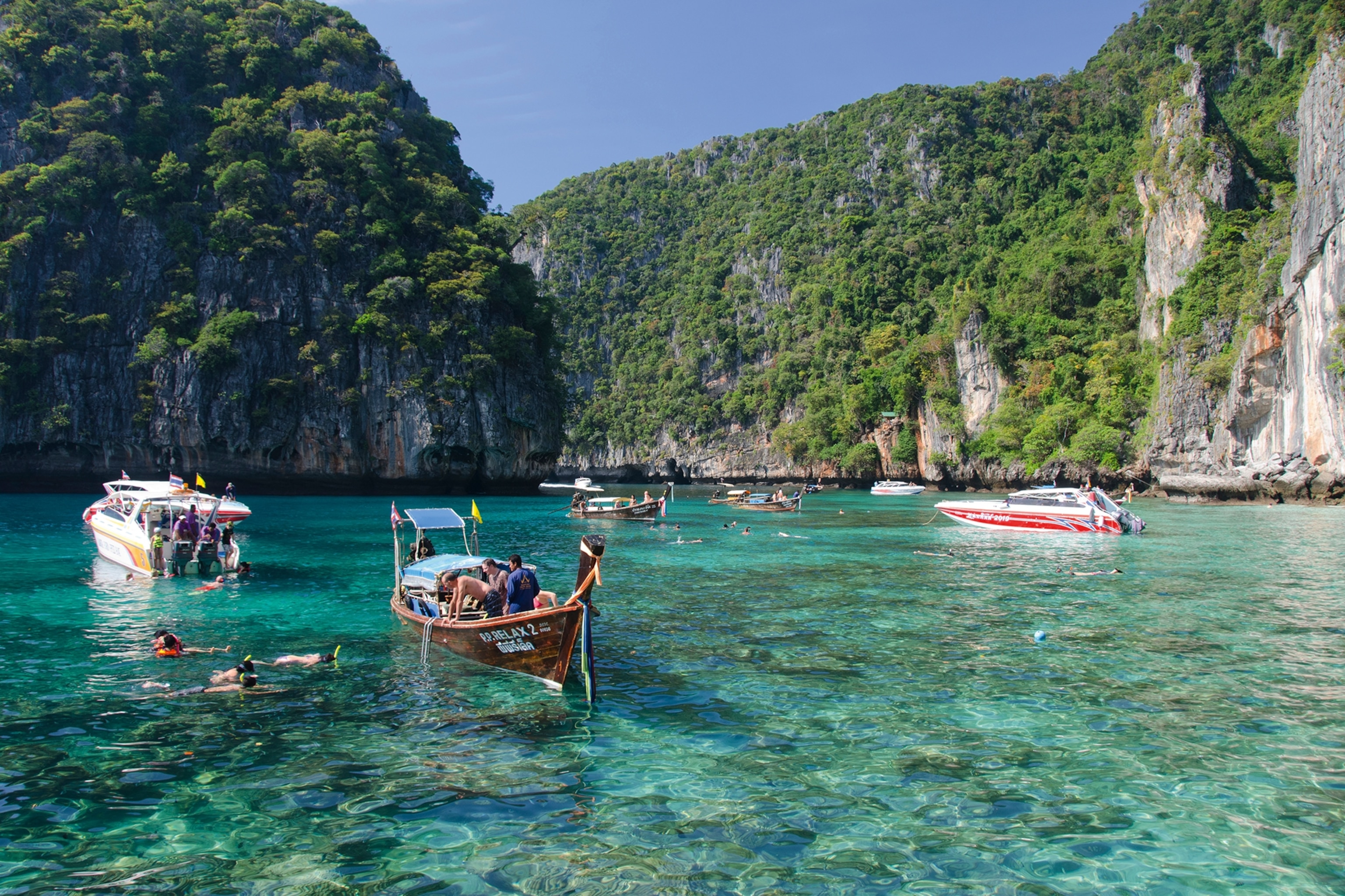 A selection of small boats surrounded by green cliffs, anchored in crystal-clear waters to snorkel.